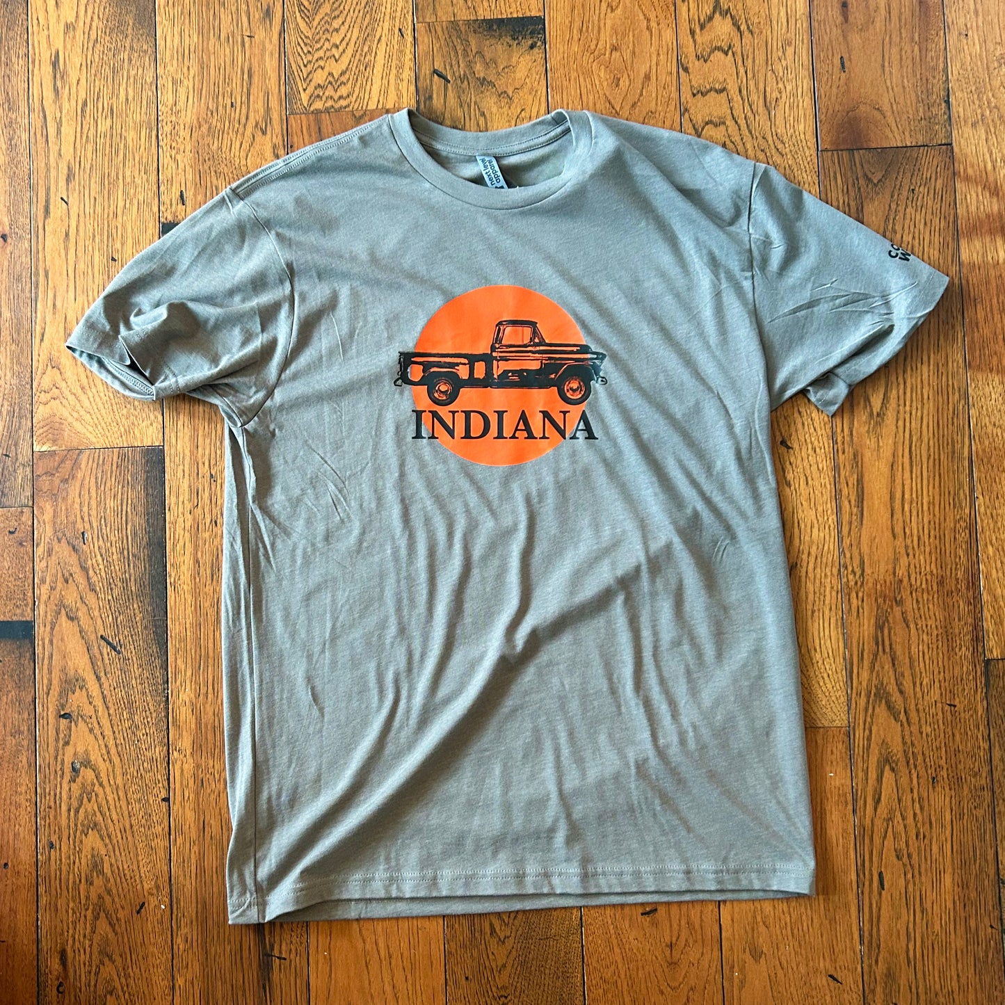 Indiana t-shirt featuring black 1956 chevy truck art on orange circle that says indiana on gray shortnsleeve t-shirt on wood flor background