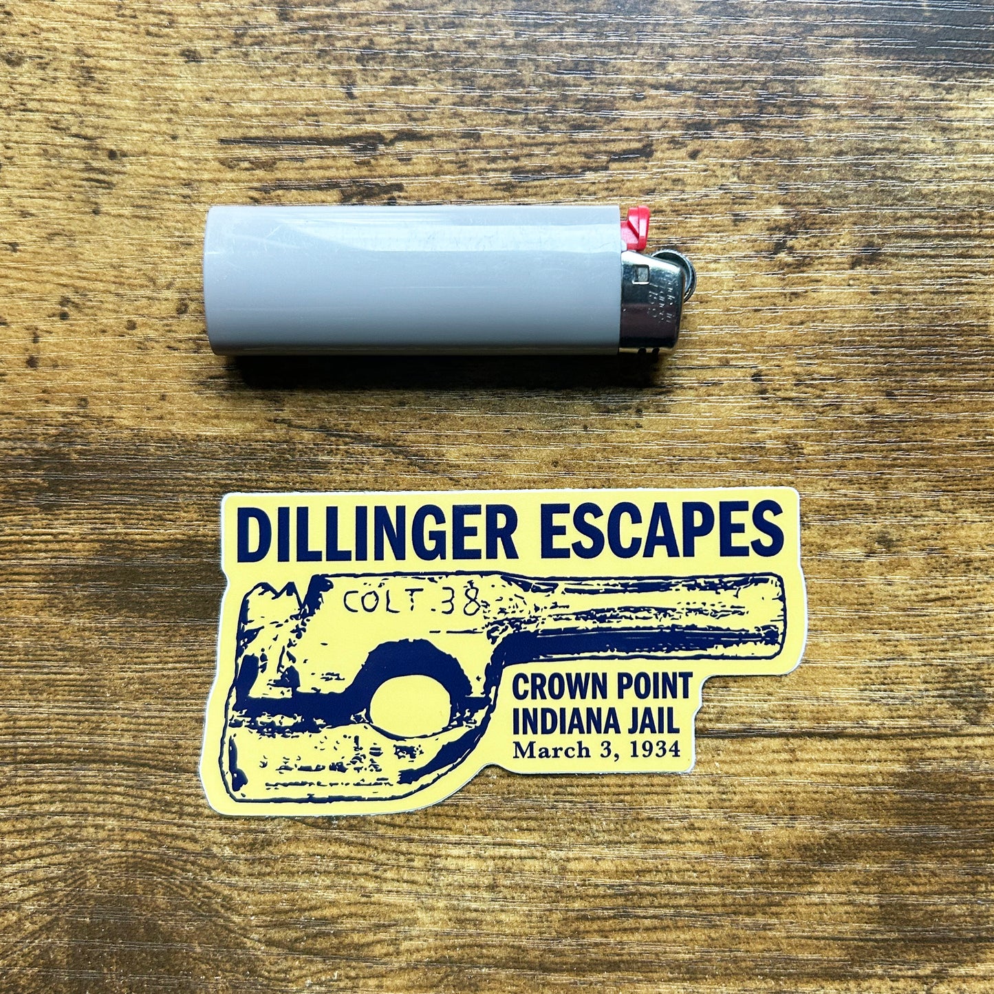 John Dillinger Escapes Crown Point Indiana Jail Sticker with wood gun artwork navy blue and yellow sticker says "Dillinger Escapes Crown Point Indiana Jail March 3, 1934" shown on wood background