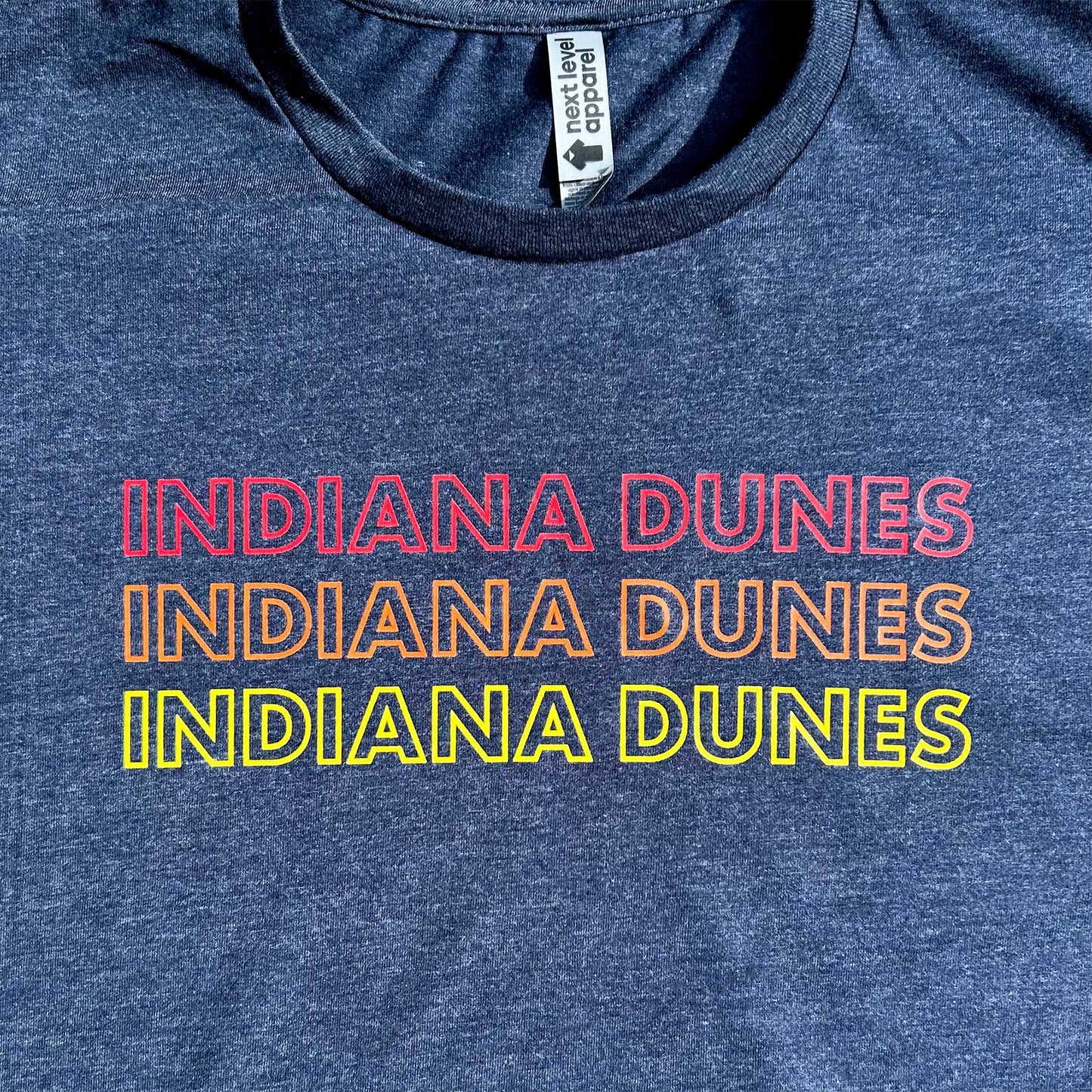 Indiana Dunes National Park T-shirt says Indiana Dunes 3 times in red, orange and yellow letters all capital letter bold outline font on navy blue shirt