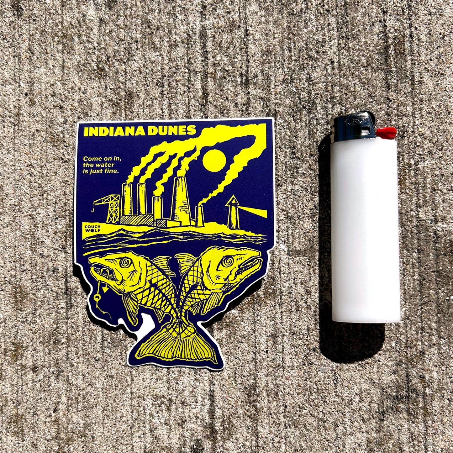 Sticker with industrial design and text 'Indiana Dunes' on a textured surface next to a lighter.