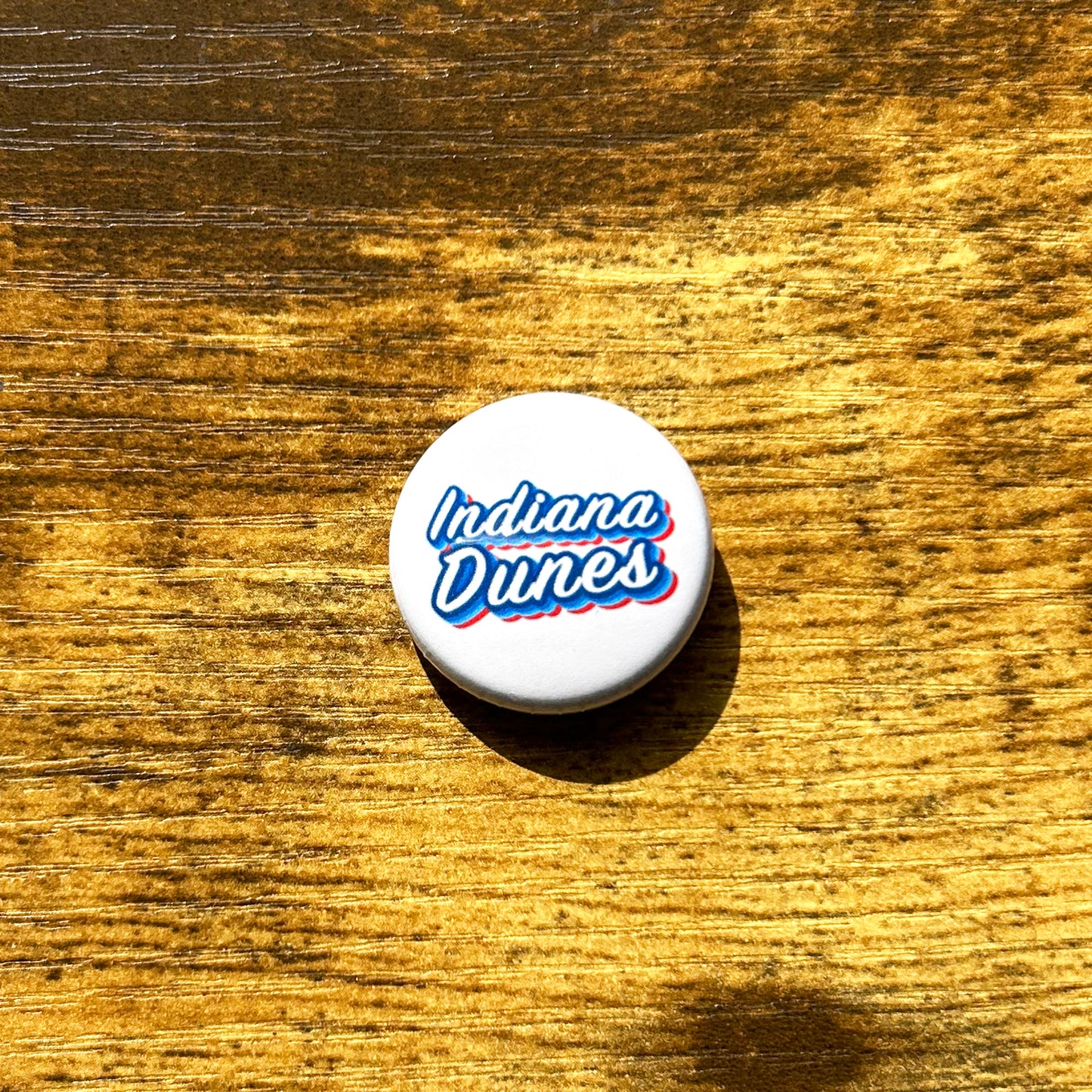Indiana Dunes pin white round pin on wood table says Indiana dunesin blue and red letters