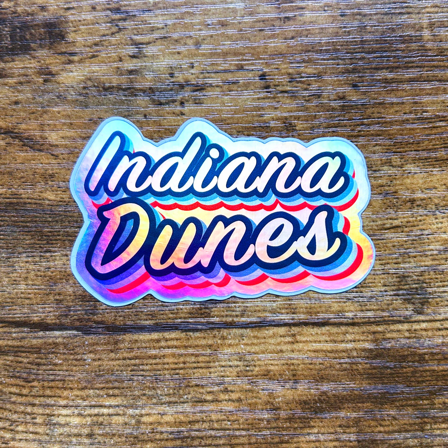 Indiana Dunes Hologram Sticker, shiny multi color reflective sticker on wood surface says "Indiana Dunes"