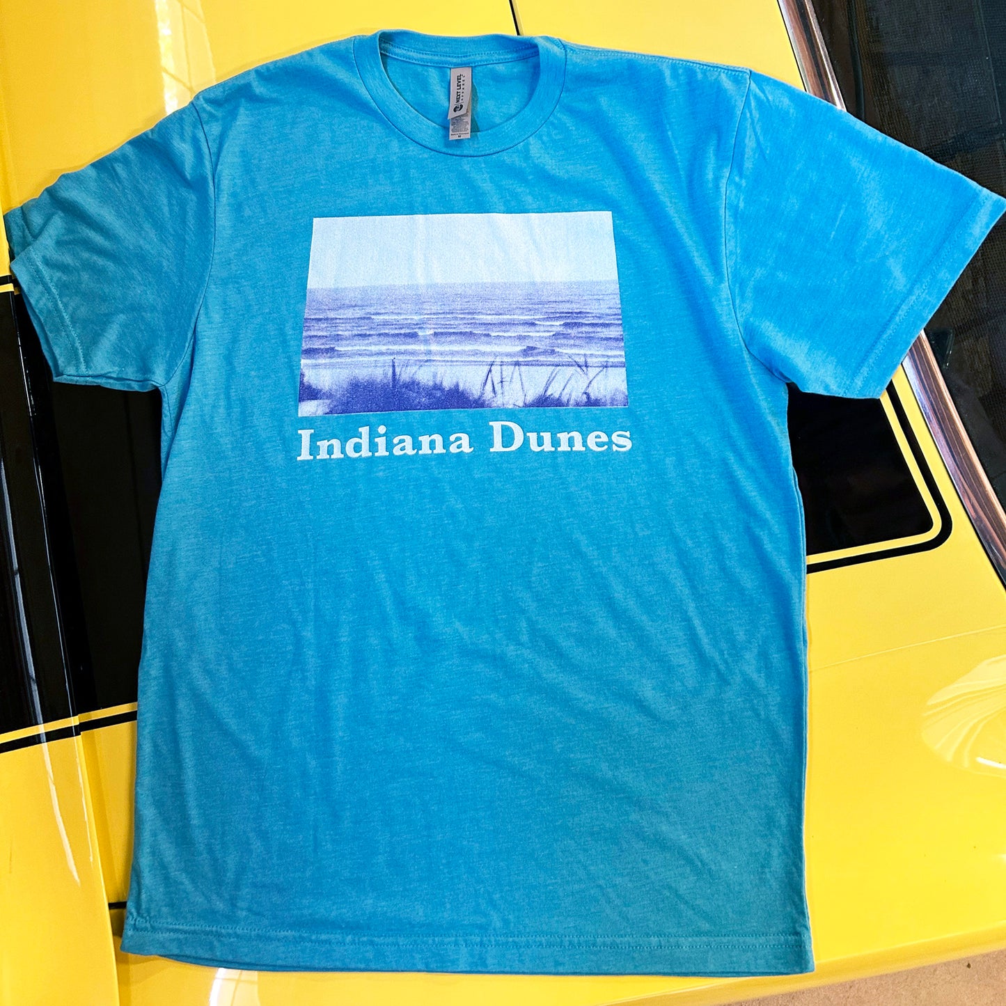 Indiana Dunes Shirt Indiana Dunes Photography t-shirt bright blue heather shirt with bright blue duotone photo of indiana Dunes National Park beach descending into lake michigan shirt on yellow car withh black stripe background
