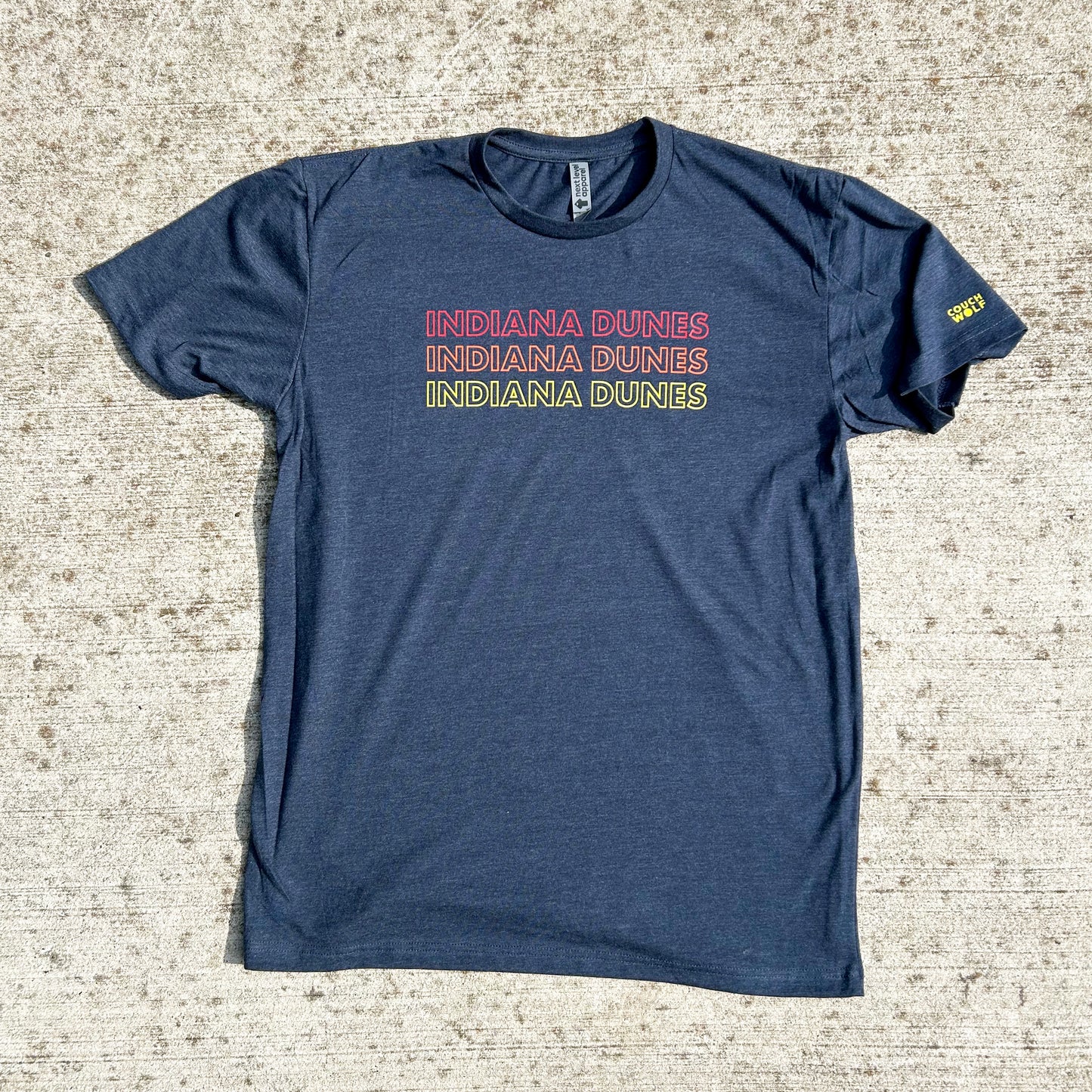 Indiana Dunes National Park T-shirt says Indiana Dunes 3 times in red, orange and yellow letters all capital letter bold outline font on navy shirt on concrete spotted background yellow couch wolf logo on sleeve