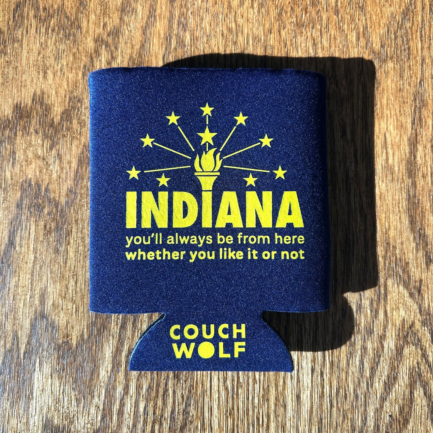 Dark blue can cooler with yellow text and 'Couch Wolf' branding on a wooden surface says "Indiana you'll always be from here whether you like it or not."