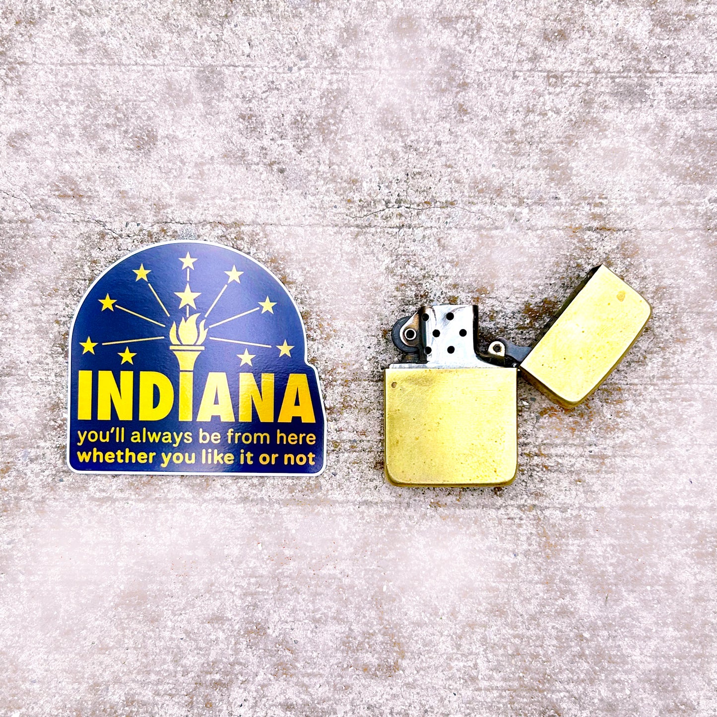 Indiana Hologram Sticker says Indiana you'll always be from here whether you like it or not in gold reflective letters on blue background with Indiana torch and stars flag shown on concrete background next to gold zippo lighter