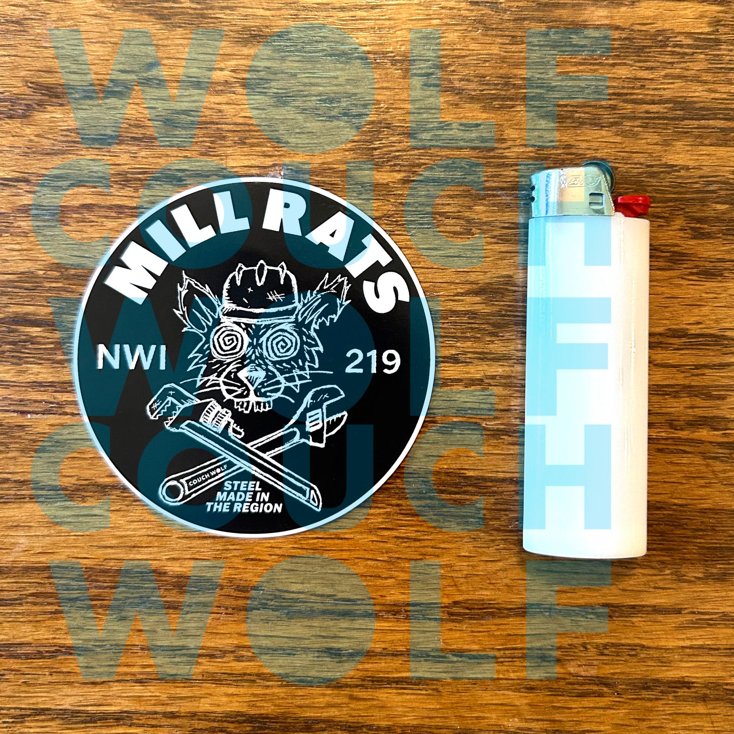 A circular black and blue sticker featuring a rat in a hard hat and crossed tools, with the text 'MILL RATS' and 'NWI 219', shown next to a white lighter on a wooden background.