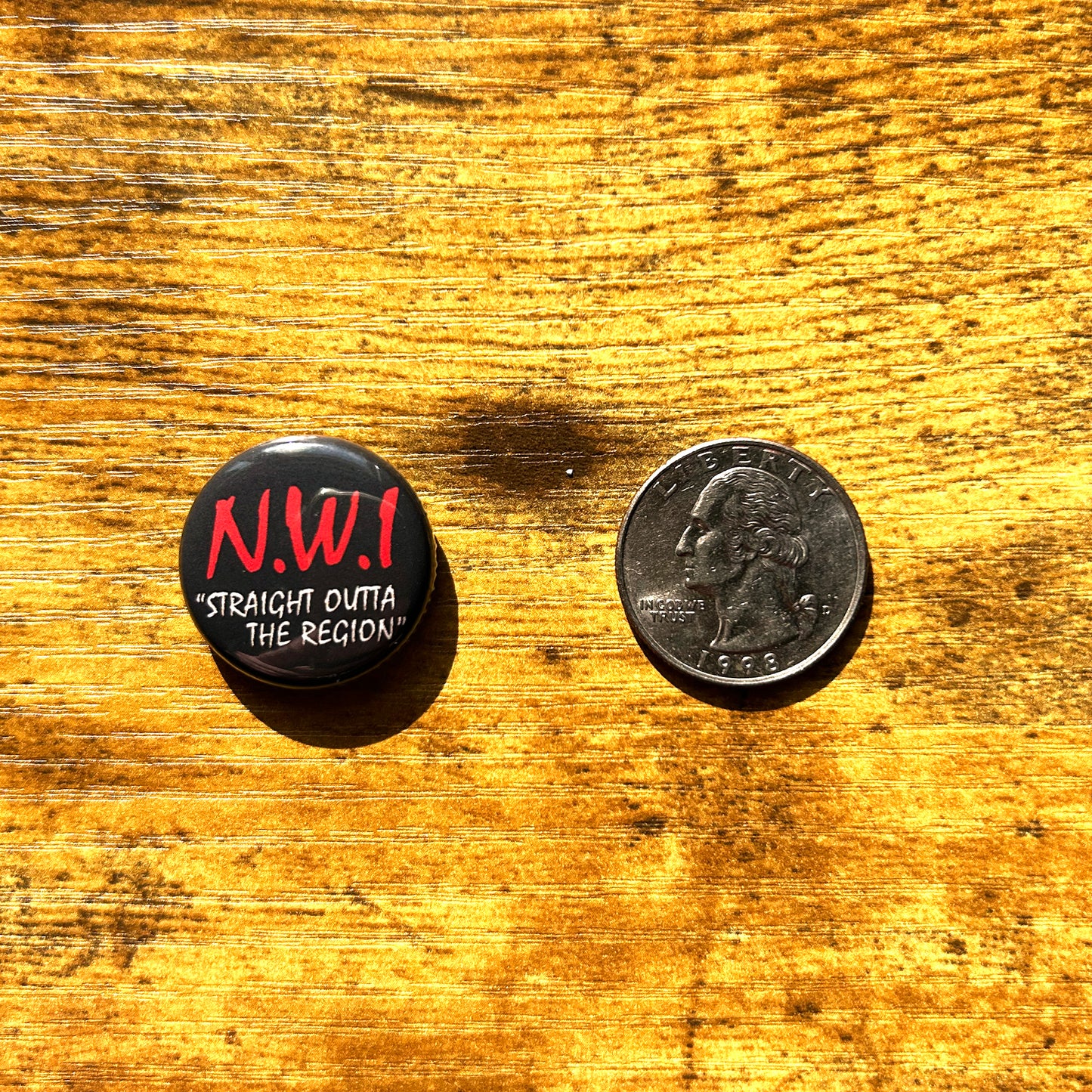 Black circle pin that says NWI straight outta the Region next to quarter on wood surface