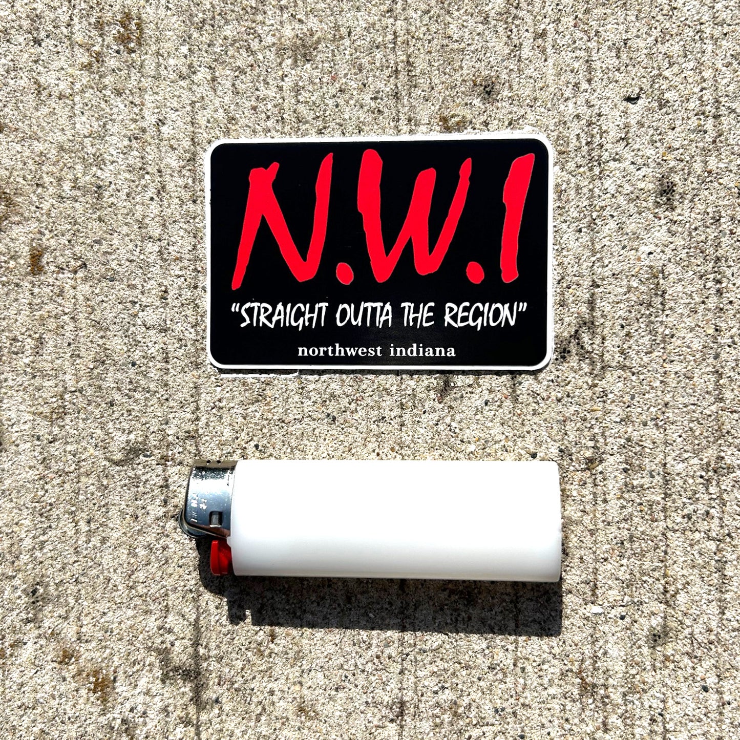 Sticker with 'N.W.I. Straight Outta The Region' text on a concrete surface with a lighter.