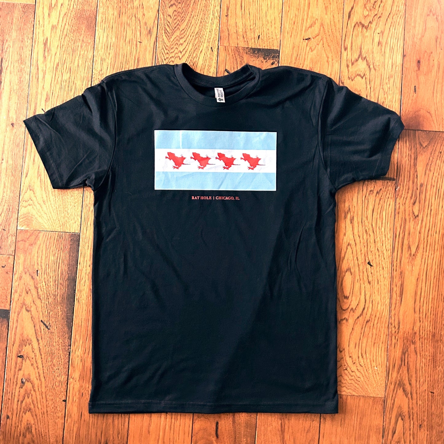 Rat Hole Chicago Flag Shirt blue and white stripe flag with red rat imprints instead of stars says "Rat Hole Chicago, IL" in red capital letters black crew neck short sleeve shirt on wood floor background.