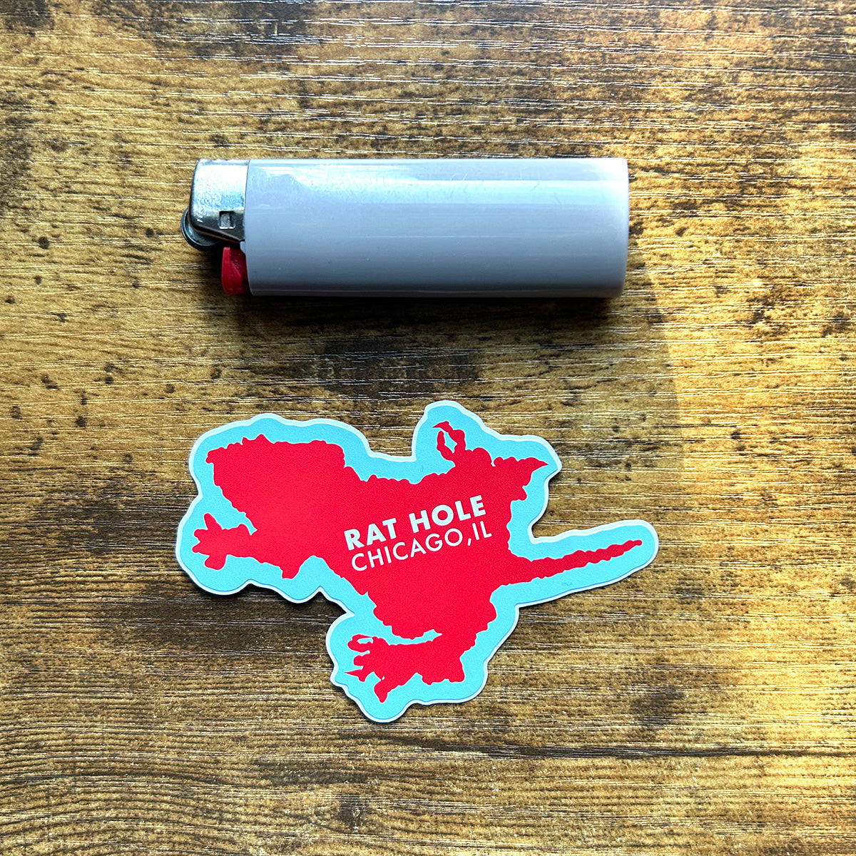 Chicago Rat Hole Sticker red and blue sticker in the shape of rat hole says rat hole chicago, il in white letters on wood table next to gray bic lighter