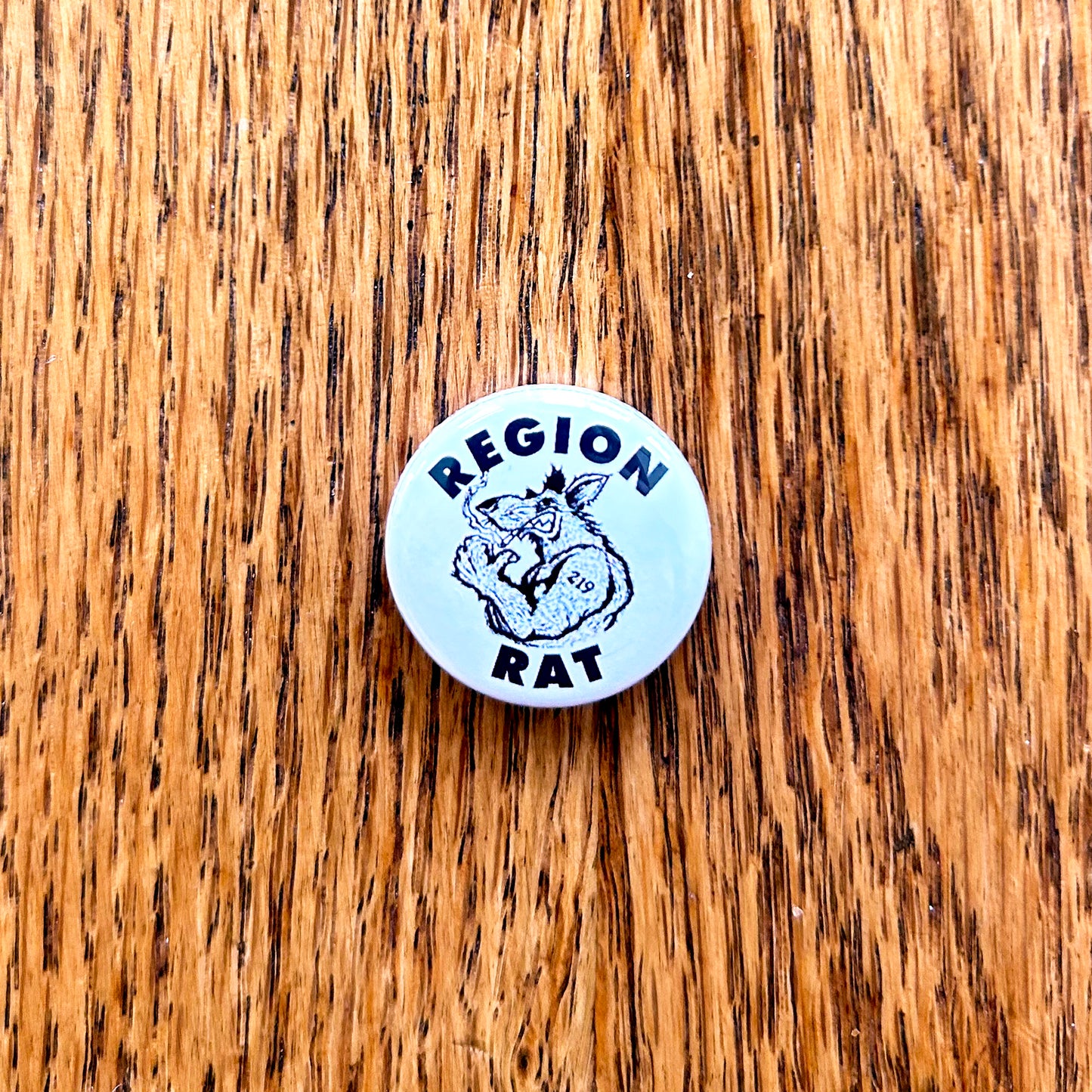 A circular pin with a hand-drawn cartoon of a rat flexing with the text 'REGION RAT' on it, placed on a wooden surface.