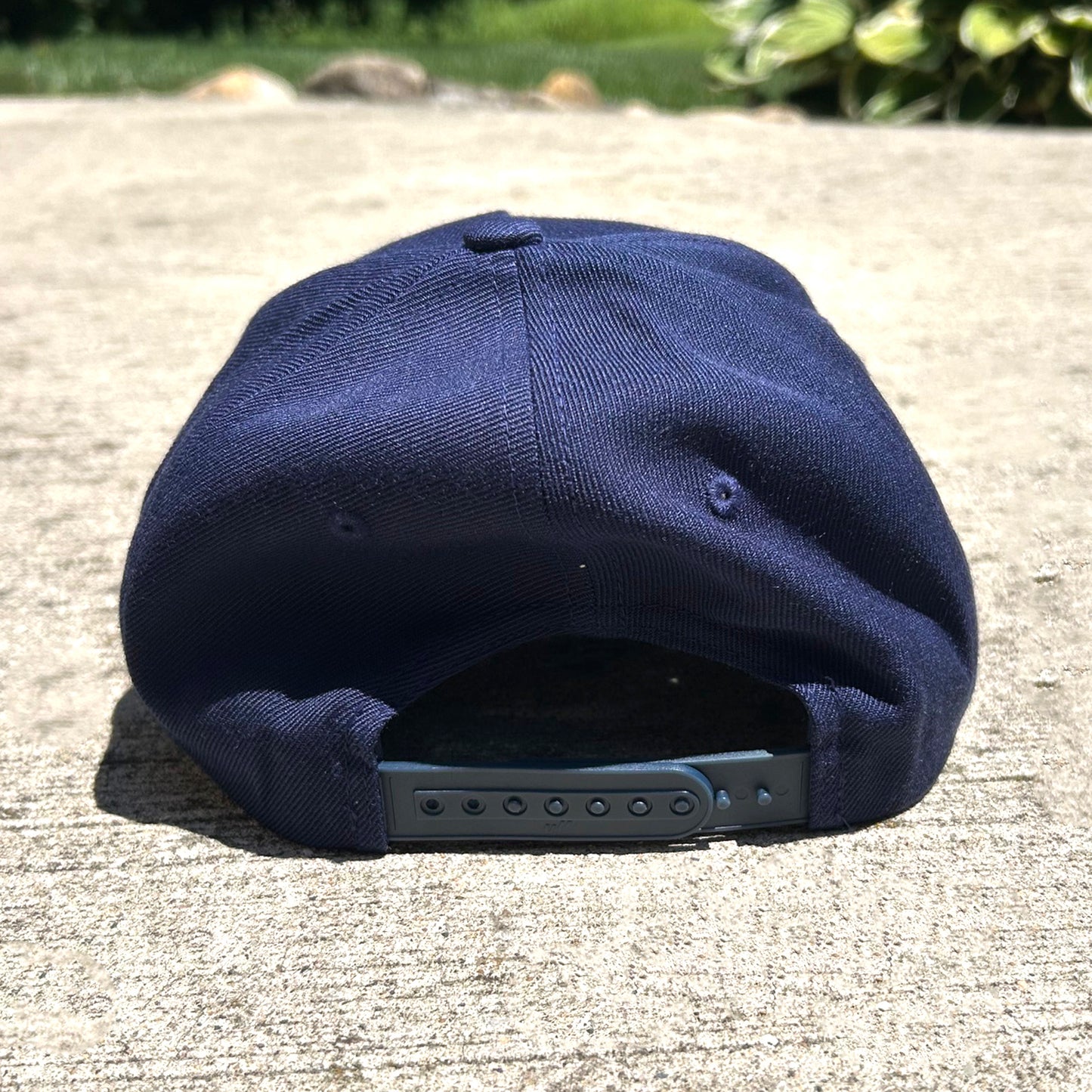 A navy blue baseball cap viewed from the back, resting on a concrete surface outdoors with greenery in the background.