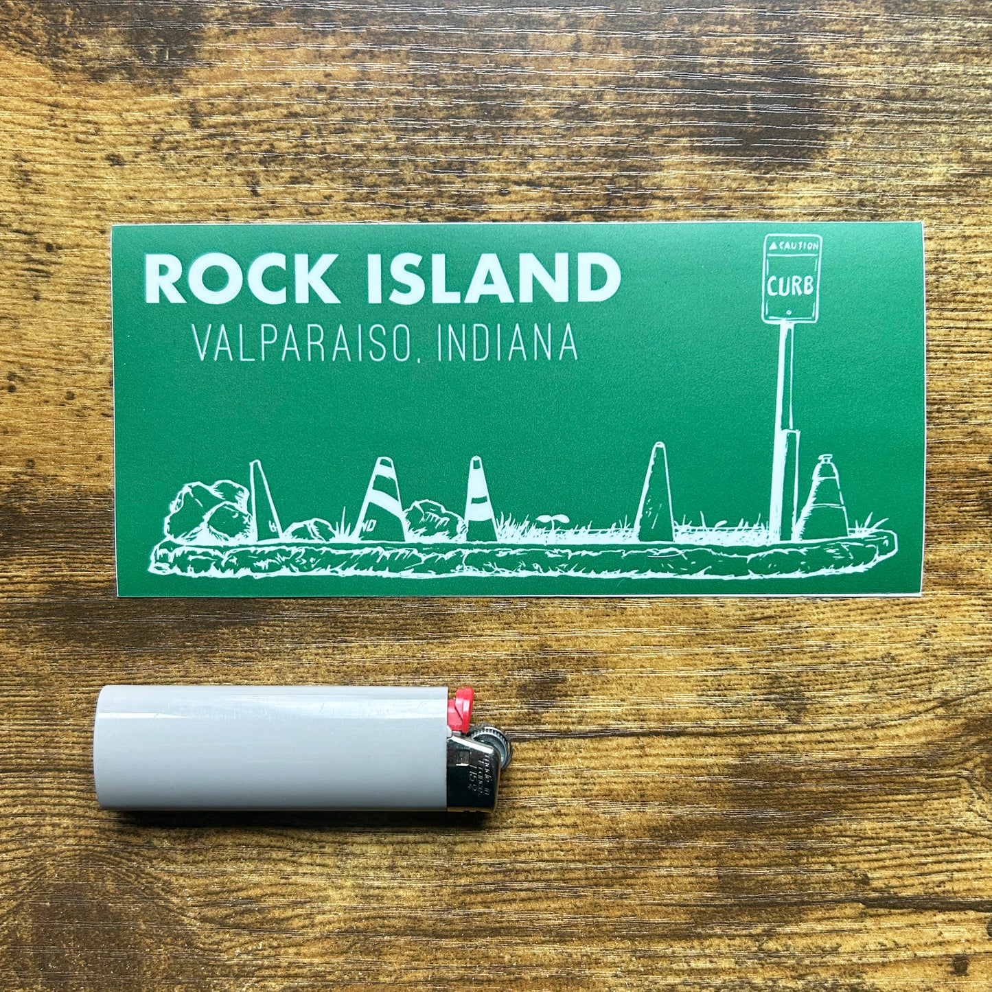 Rock Island Valparaiso Indiana Sticker green sticker with white line art of parking island rocks and road cones says "Rock Island Valparaiso Indiana" shown on wood background next to gray bic lighter