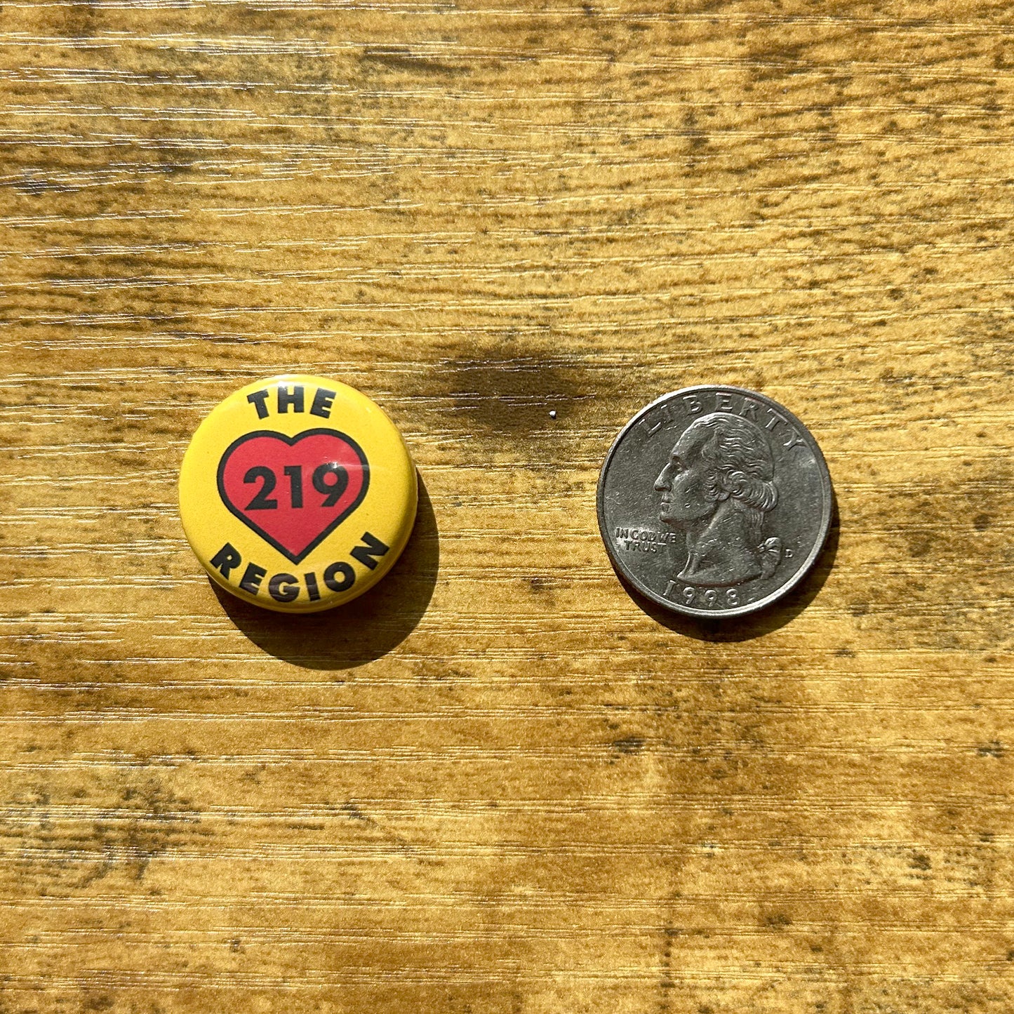 The Region 219 heart pin yellow pin with black letter that say the region 219, 219 is inside of red heart, circle pin shown on wood grain table next to us quarter for scan