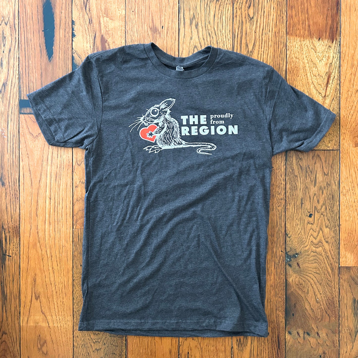 Region Rat Indiana shirts says proudly from the region with white ink draawing of rat holding red heart on gray shirt on wood floor background