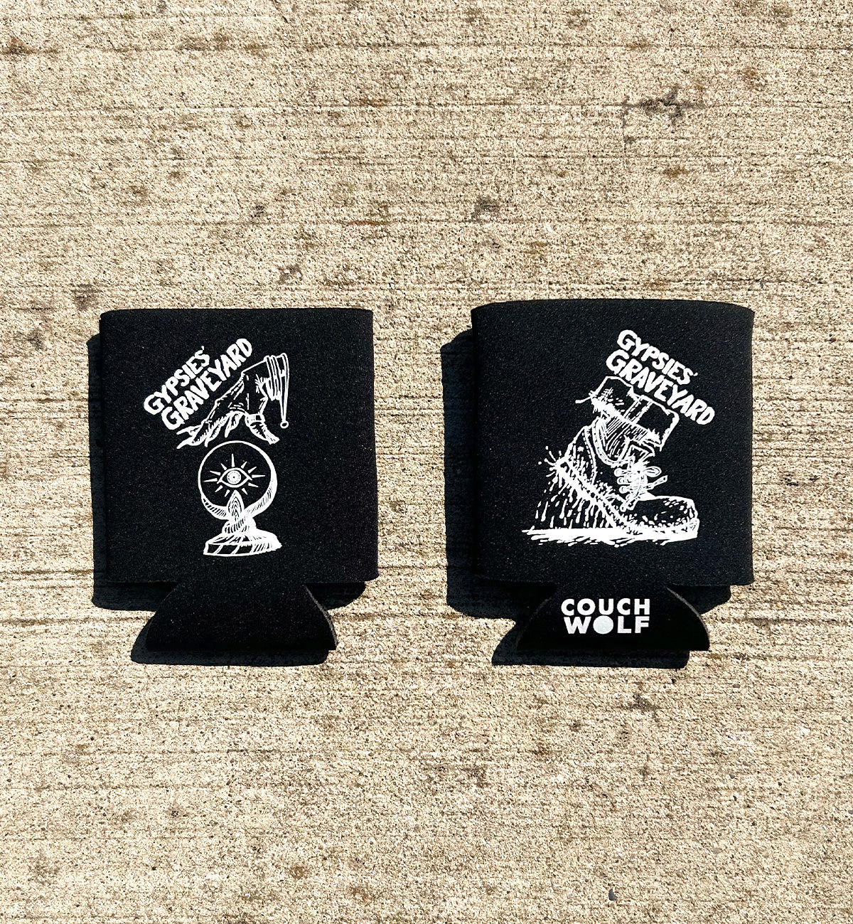 2 gypsies graveyard black can coolers foam can cooler have white ink artwork of bloody shoe stepping and hand over crystal ball and say "gypsie's graveyard" on both sides, shown on concrete background, laying flat with white couch wolf logo on bottom of show side