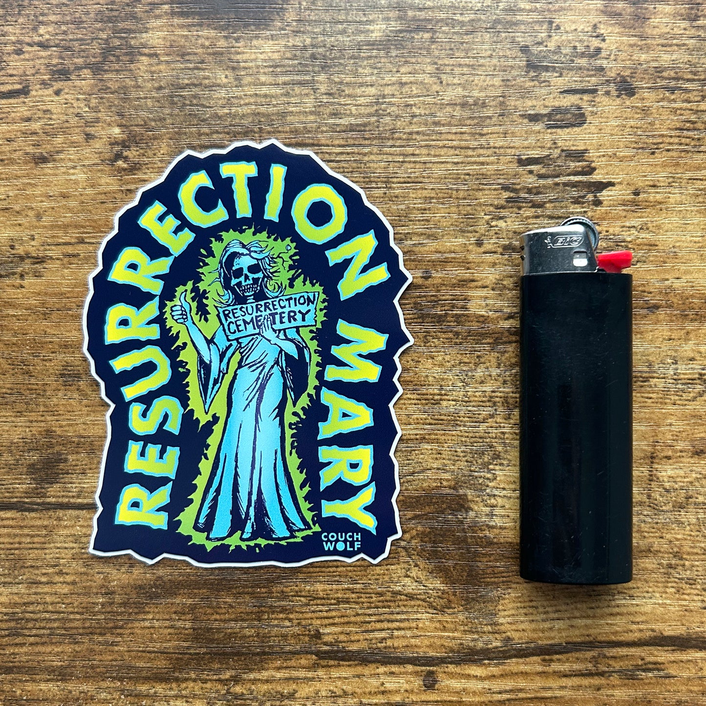 A die-cut sticker featuring a ghost holding a sign that reads 'RESURRECTION CEMETERY' with the words 'RESURRECTION MARY' curving around the top and bottom, placed on a rustic wooden surface next to a black lighter.
