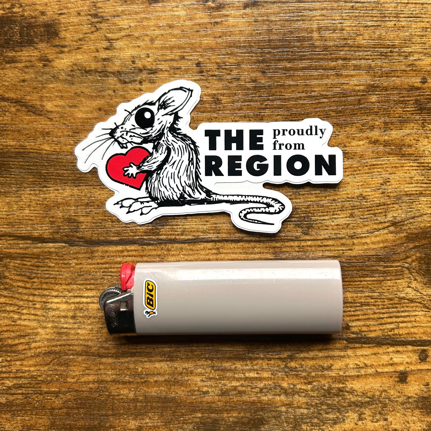 The Region rat sticker says proudly from the region in black letters next to black line art drawing of rat holding red heart on wood background next to gray pic lighter