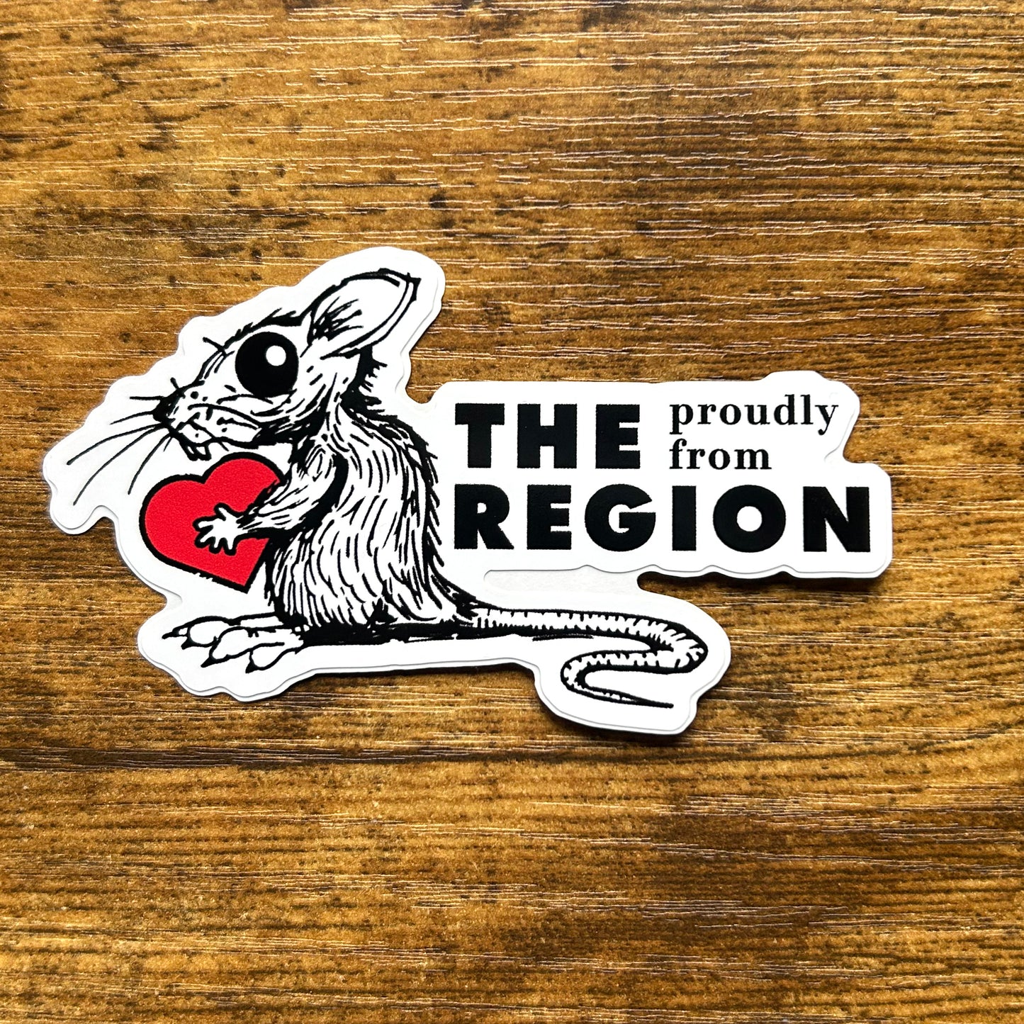 The Region rat sticker says proudly from the region in black letters next to black line art drawing of rat holding red heart on wood background