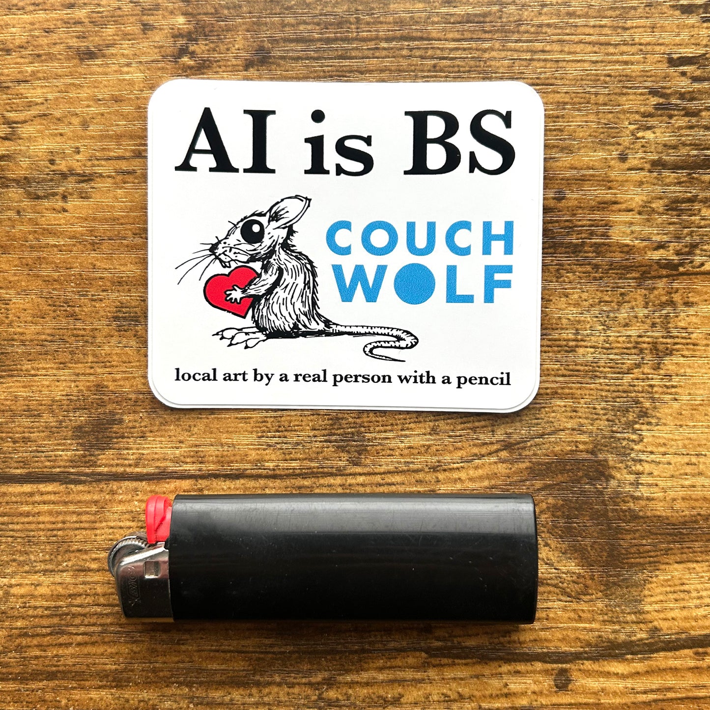 A square white sticker with the text 'AI is BS' and 'COUCH WOLF' in blue, plus an illustration of a black rat holding a red heart, placed on a wooden surface above a black lighter.