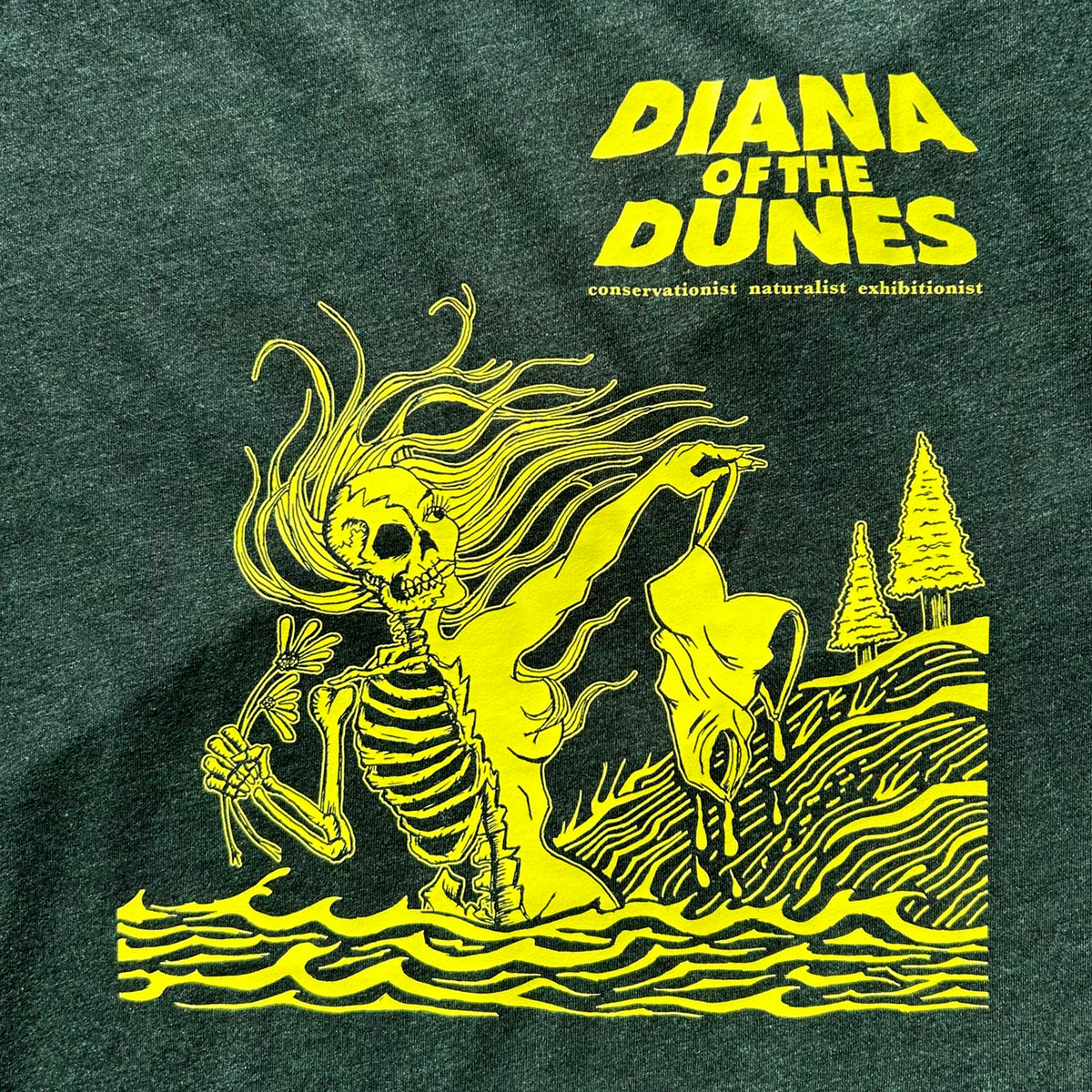 Dark heather green t-shirt featuring a bright yellow graphic print of a skeleton in a beach landscape dangling a swimsuit with the text 'DIANA OF THE DUNES' and a subtitle 'consevationist naturalist exhibitionist'.
