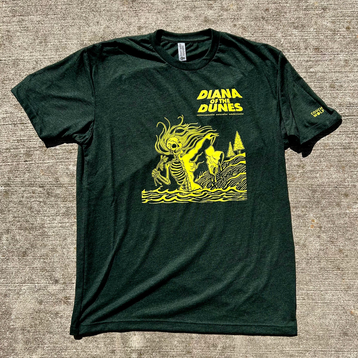 A dark forest green t-shirt featuring a yellow graphic of a skeleton swimming near a sand dune with the text 'DIANA OF THE DUNES' on the chest, laid flat on a textured gray concrete background.