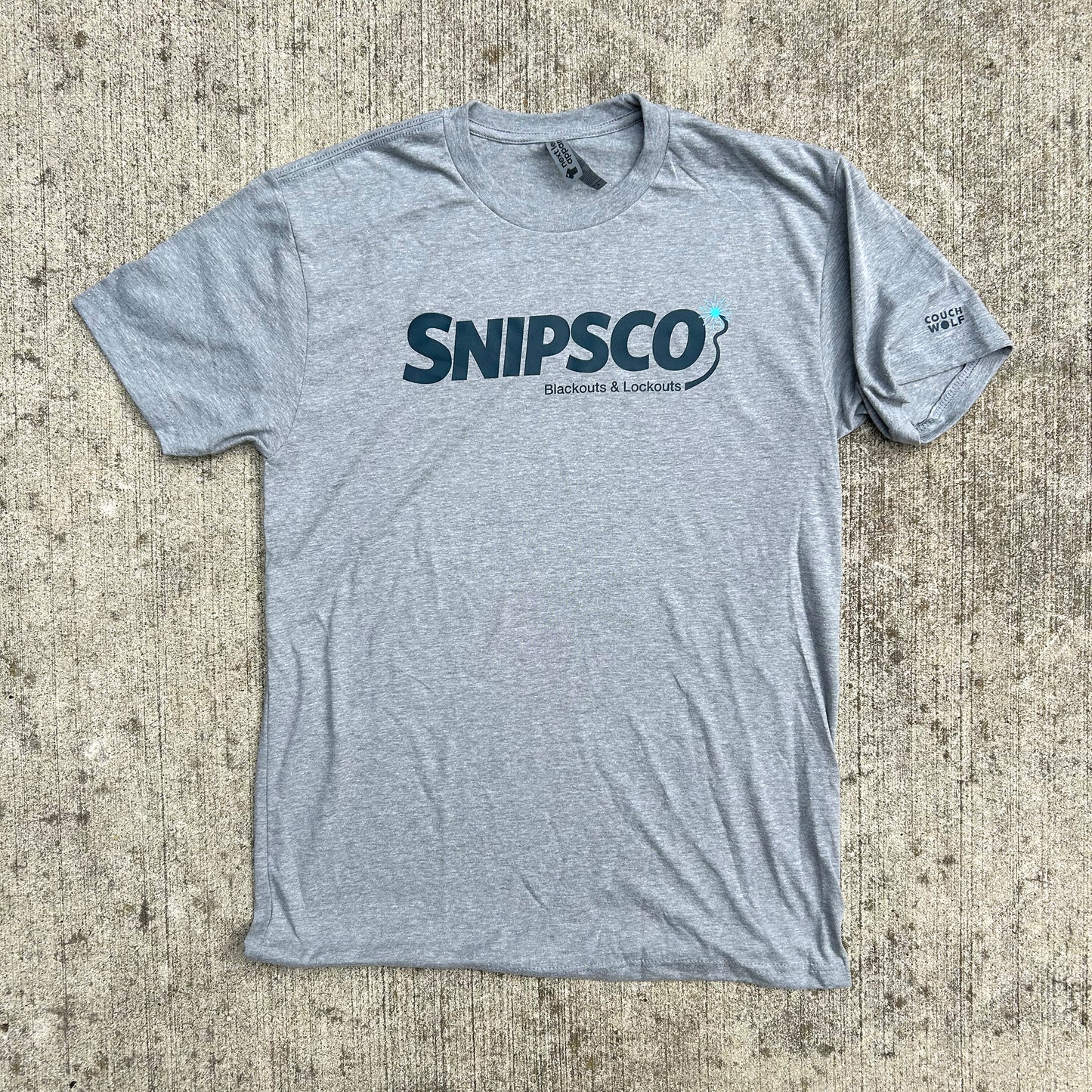 A heather gray short-sleeve t-shirt with dark navy blue 'snips-co blackouts & lockouts' graphic printed across the chest, laid flat on a textured concrete surface.