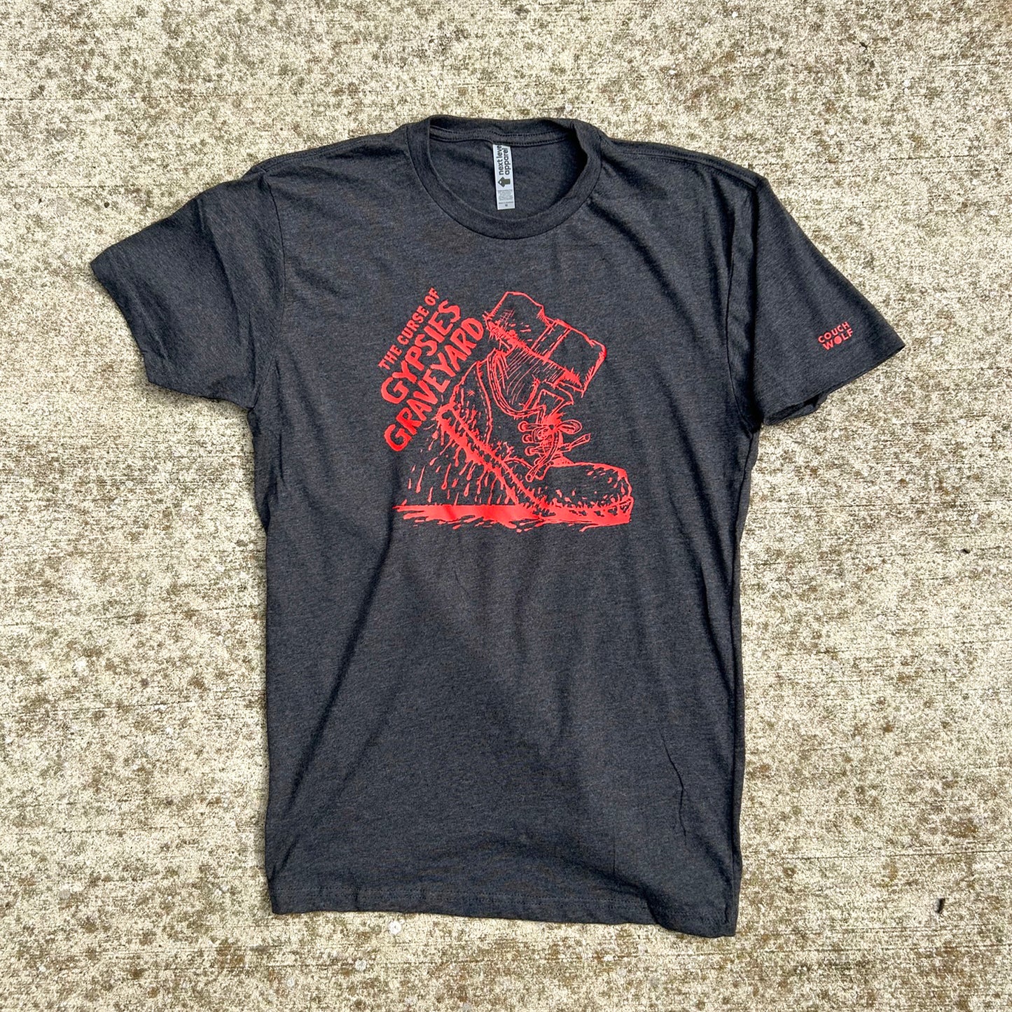 Dark heather gray t-shirt featuring a bold red graphic print of a shoe dripping blood and the text 'THE CURSE OF GYPSYS GRAVEYARD' shown on tan speckled concrete background.