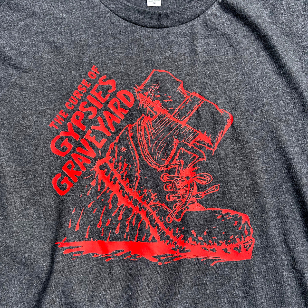 Close-up of a dark heather gray t-shirt featuring a bold red graphic print of a shoe dripping blood and the text 'THE CURSE OF GYPSYS GRAVEYARD'.