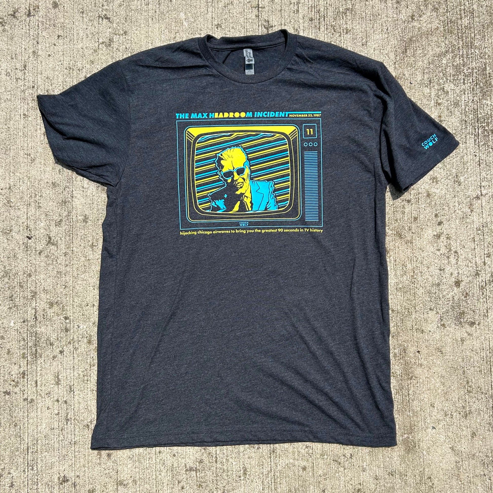 A dark heather gray graphic t-shirt laid flat on a textured concrete surface. The shirt features a prominent retro television graphic in yellowish green, blue  on the chest, depicting a figure with yellow skin and hair.