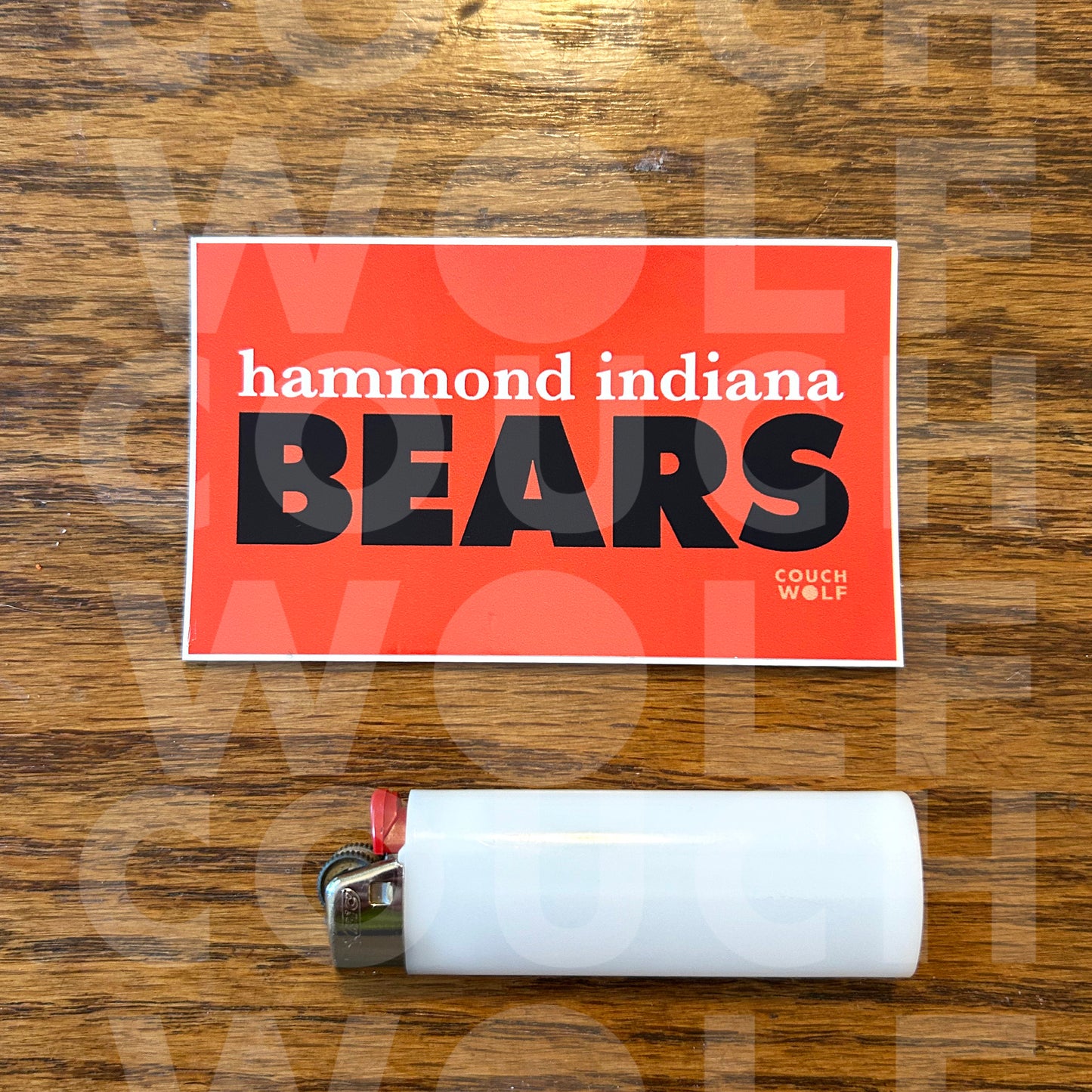 A rectangular orange sticker with the text 'hammond indiana BEARS' in white and blue, placed on a wooden surface above a white disposable lighter.
