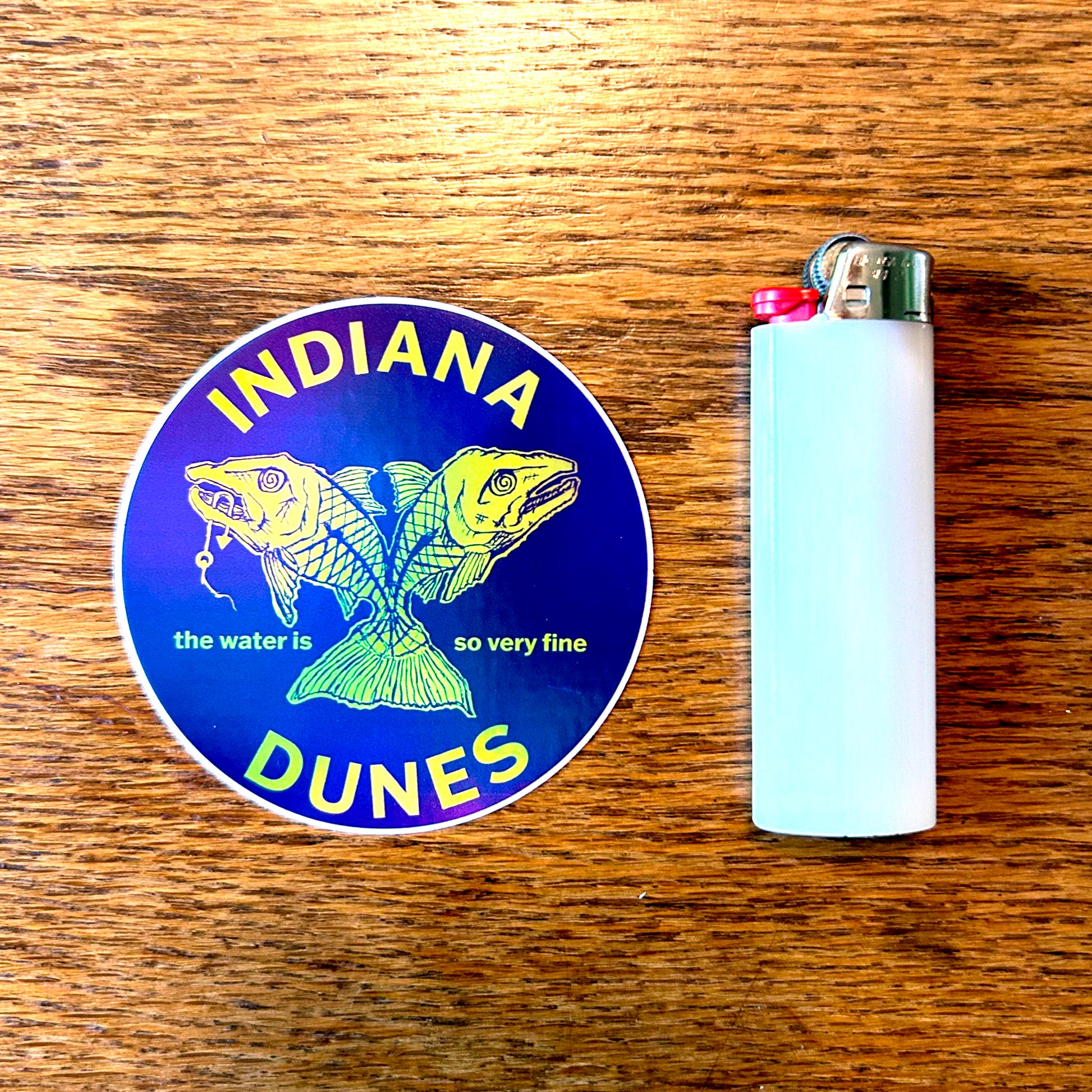 Indiana Dunes sticker with two fish illustrations on a blue background, next to a lighter on a wooden surface.