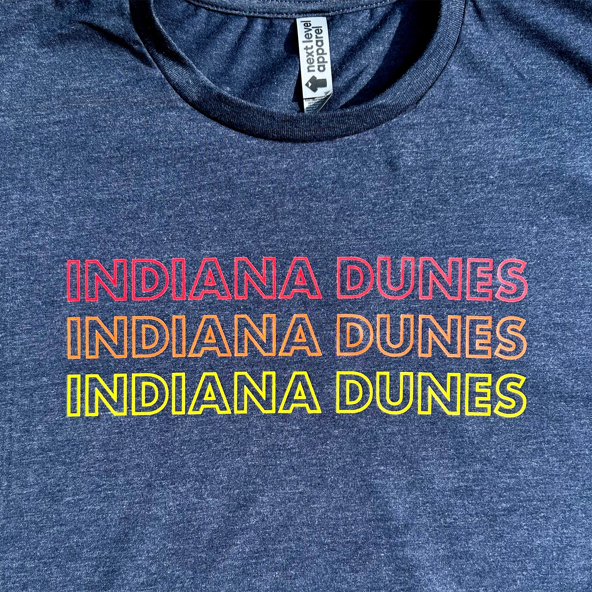 Indiana Dunes National Park T-shirt says Indiana Dunes 3 times in red, orange and yellow letters all capital letter bold outline font on navy blue shirt