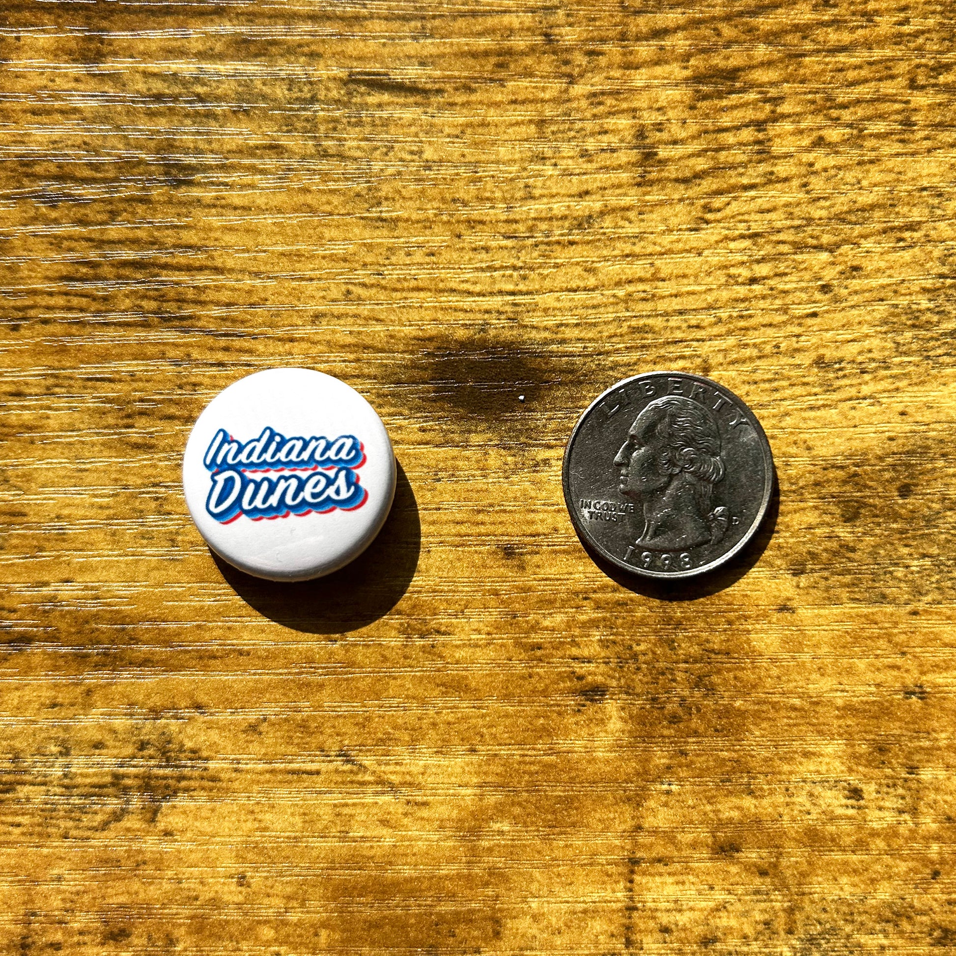 Indiana Dunes pin white round pin next US quarter for scale on wood table, pin says Indiana dunes in white letters with red and blue outlines