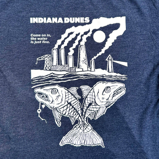 Indiana Dunes National Park T-shirt says Indiana Dunes  Ciome on in the water is just fine. White ink artwork on navy blue shirt features 2-headed fish with hook in one of it's mouths swimming beneath the smoggy sky polluted by steel mill smoke stacks  shown on concrete floor background