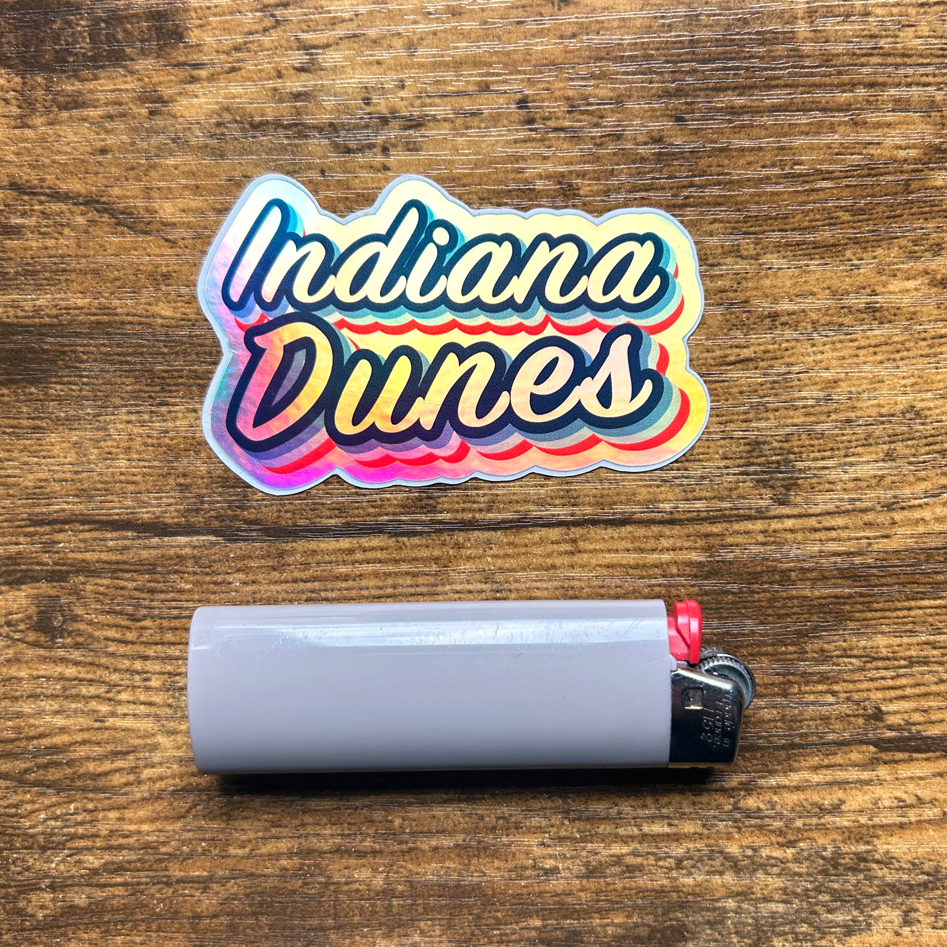 Indiana Dunes Hologram Sticker, shiny multi color reflective sticker with multiple ble inks and red ink on wood table says "Indiana Dunes" next to gray bic lighter for scale