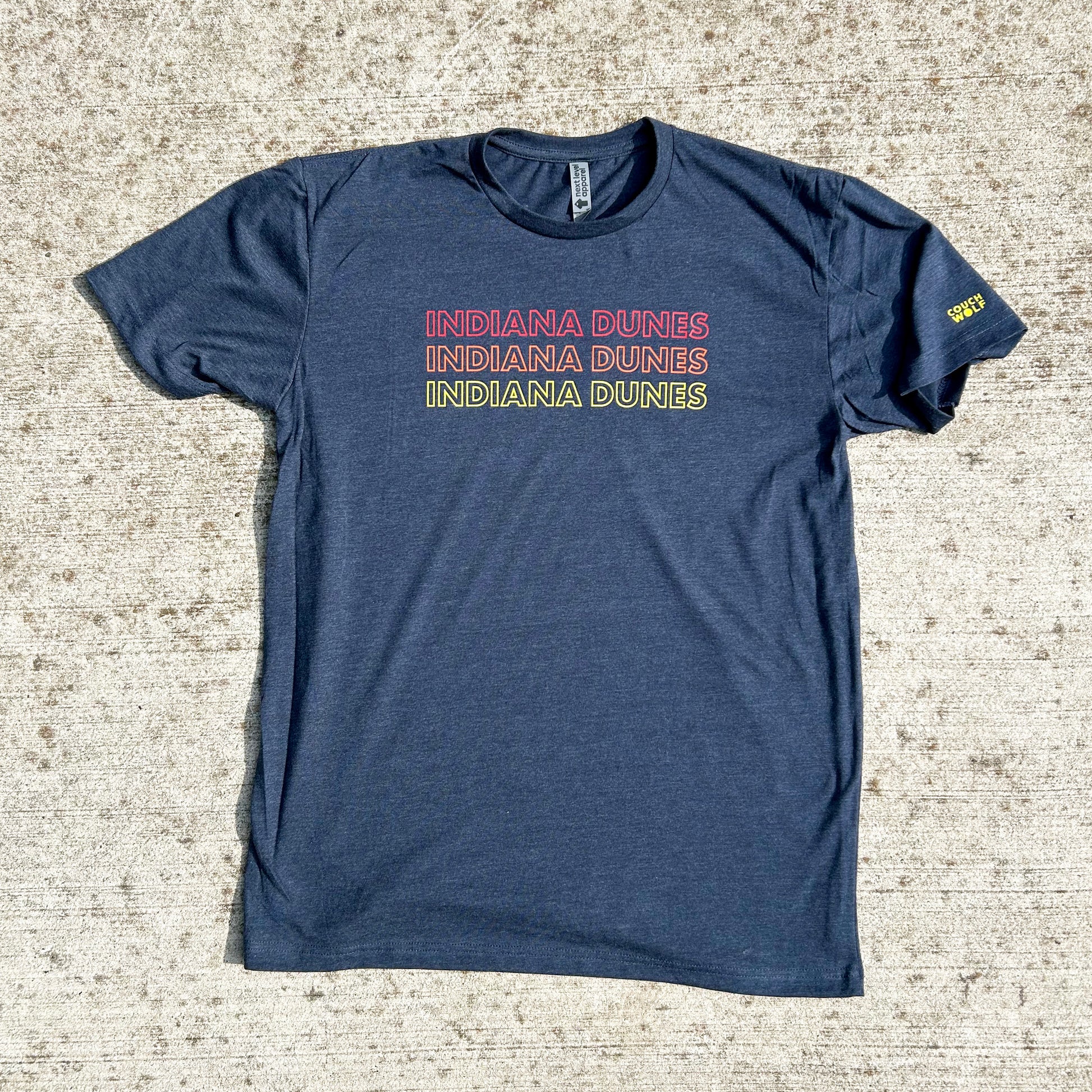 Indiana Dunes National Park T-shirt says Indiana Dunes 3 times in red, orange and yellow letters all capital letter bold outline font on navy shirt on concrete spotted background yellow couch wolf logo on sleeve