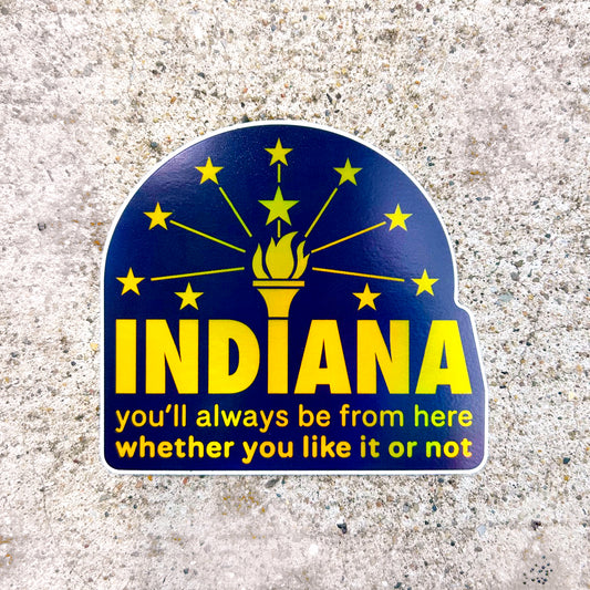 Indiana Hologram Sticker says Indiana you'll always be from here whether you like it or not in gold reflective letters on blue background with Indiana torch and stars flag shown on concrete background