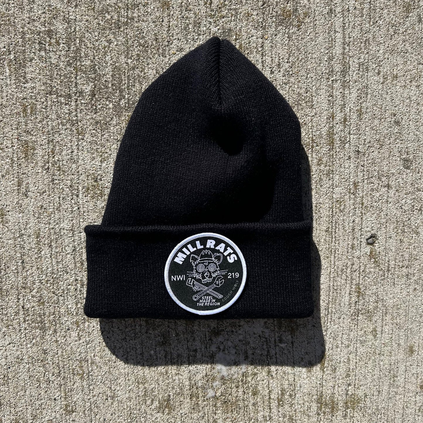 Black beanie with a Mill Rats logo patch on a concrete surface