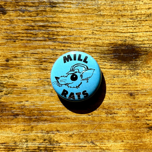 A blue pin with 'MILL RATS' text and a hand-drawn rat wearing a hard hat artwork on it, placed on a wooden surface.