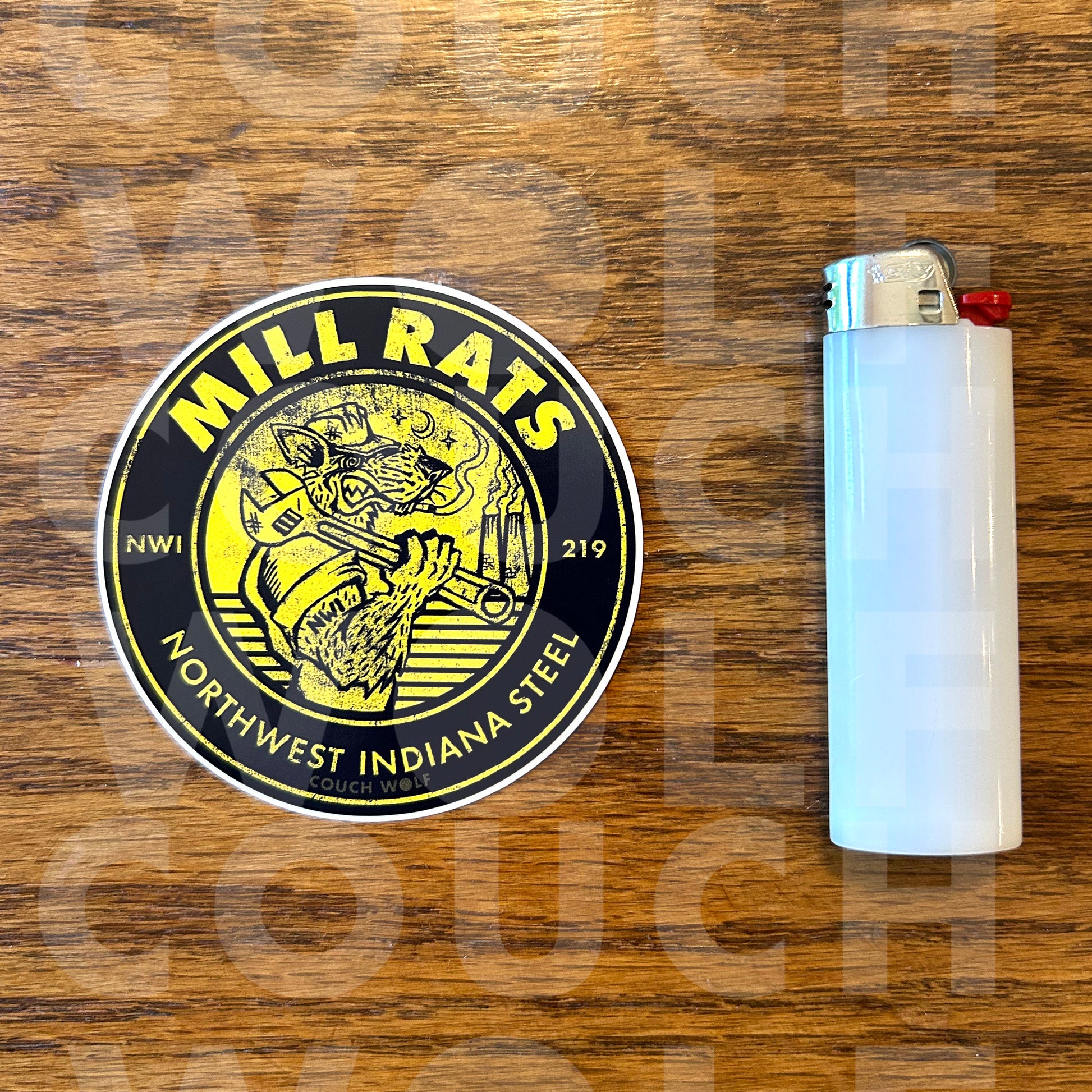A circular black and yellow 'Mill Rats Northwest Indiana Steel' sticker is shown next to a white plastic lighter on a brown wooden background.