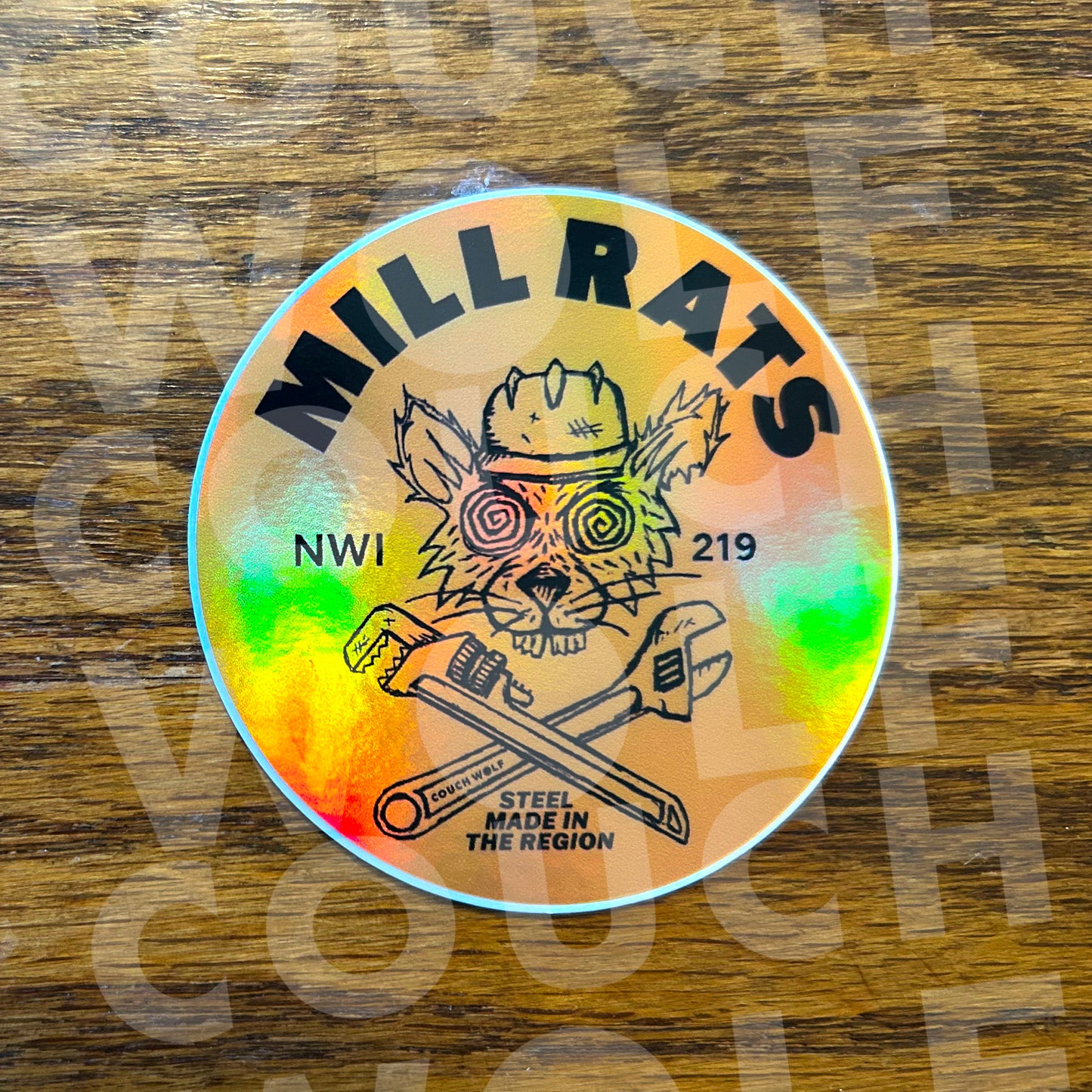 A round holographic sticker featuring a cartoon rat wearing a hard hat with the text 'MILL RATS' and 'STEEL MADE IN THE REGION', placed on a wooden surface.