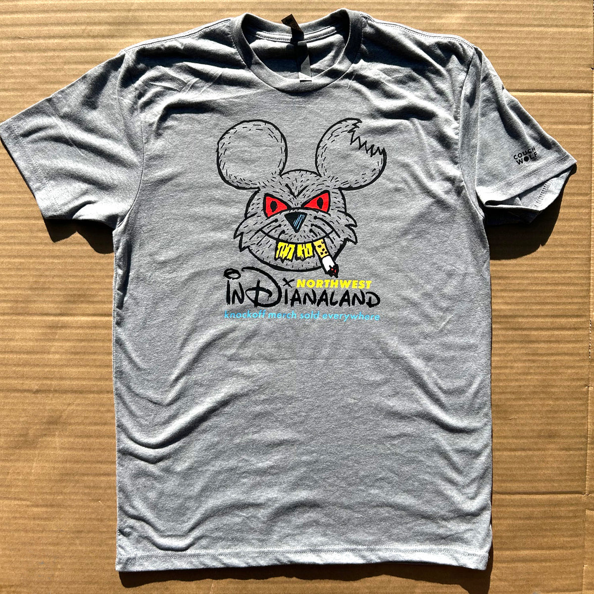 A heather gray short-sleeve t-shirt laid flat on a brown cardboard background. The shirt features a graphic of a cartoon rat head with red eyes, yellow teeth, and the text that says 'NORTHWEST INDIANALAND ' and below it in blue text it says 'knockoff merch sold everywhere."