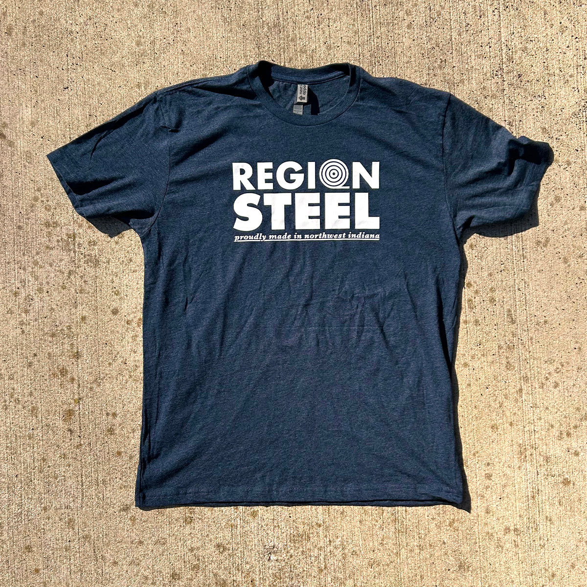 Region Steel Indiana T-shirt navy blue shirt with white bold letters that say "Region Steel proudly made in northwest Indiana, short sleeve shirt on brown concrete background