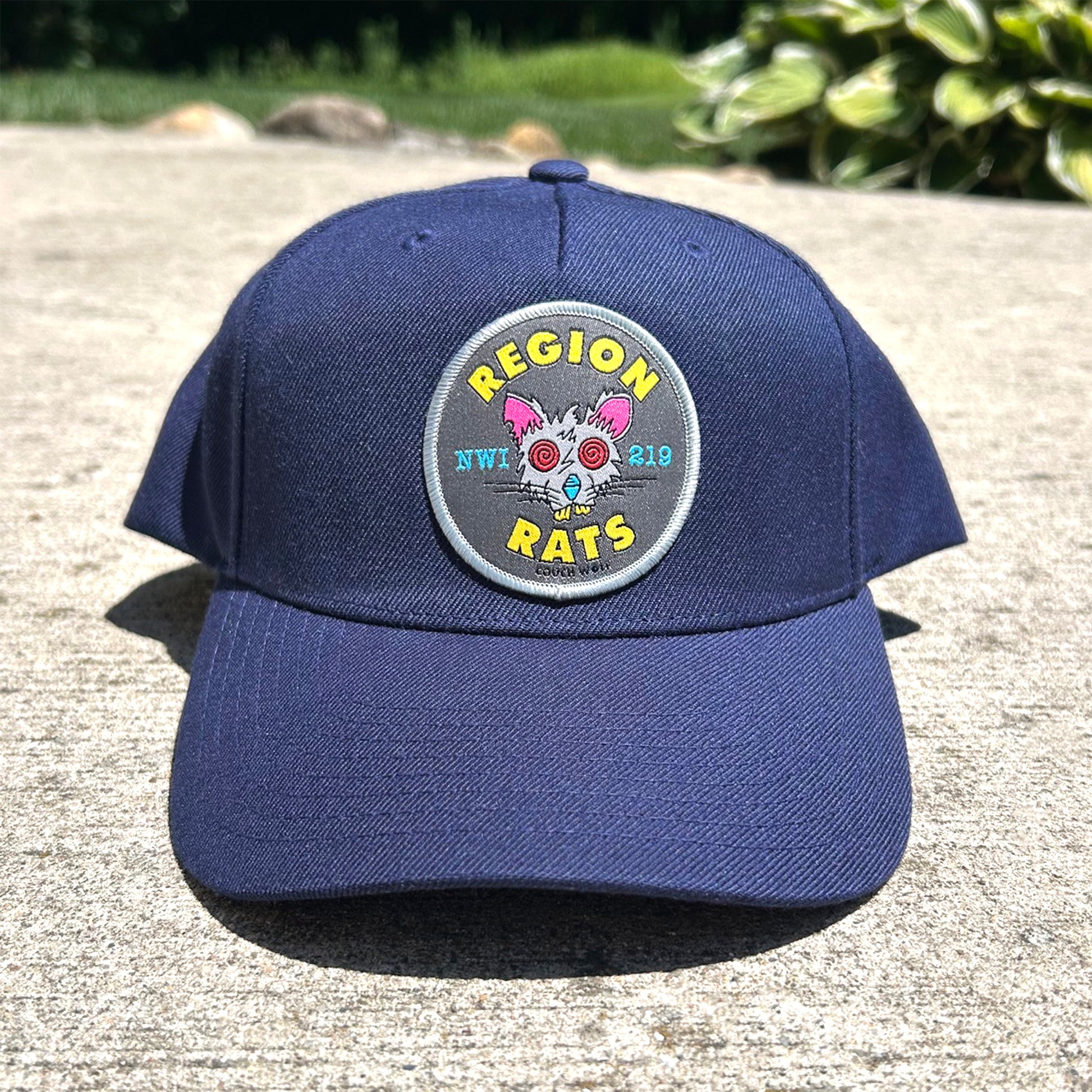 A navy blue baseball cap with a circular embroidered patch on the front that reads 'REGION 2025 RATS' and features a cartoon rat design, photographed outdoors on a concrete surface with blurred greenery in the background.