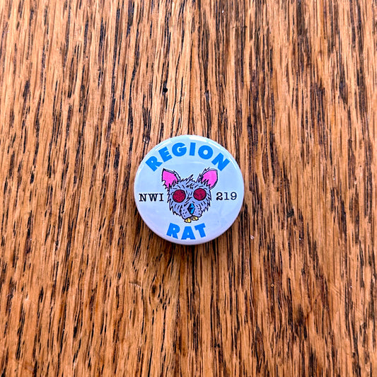 Region Rat Pin Northwest Indiana pin rat artwork features gray rat with red eyes and pink ears artwork says region rat in blue letters  nwi 219 in black on white circle pin shown on wood grain table