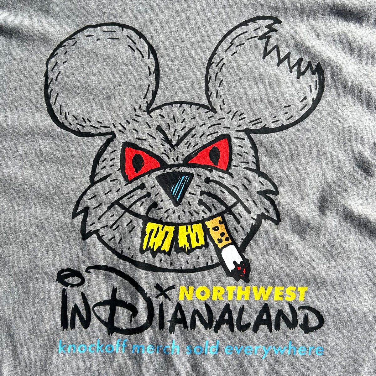 Close-up of a heather gray T-shirt featuring a stylized cartoon rat head with red eyes, a black nose, yellow text reading 'NORTHWEST INDIANALAND', and a small smoking cigarette graphic.