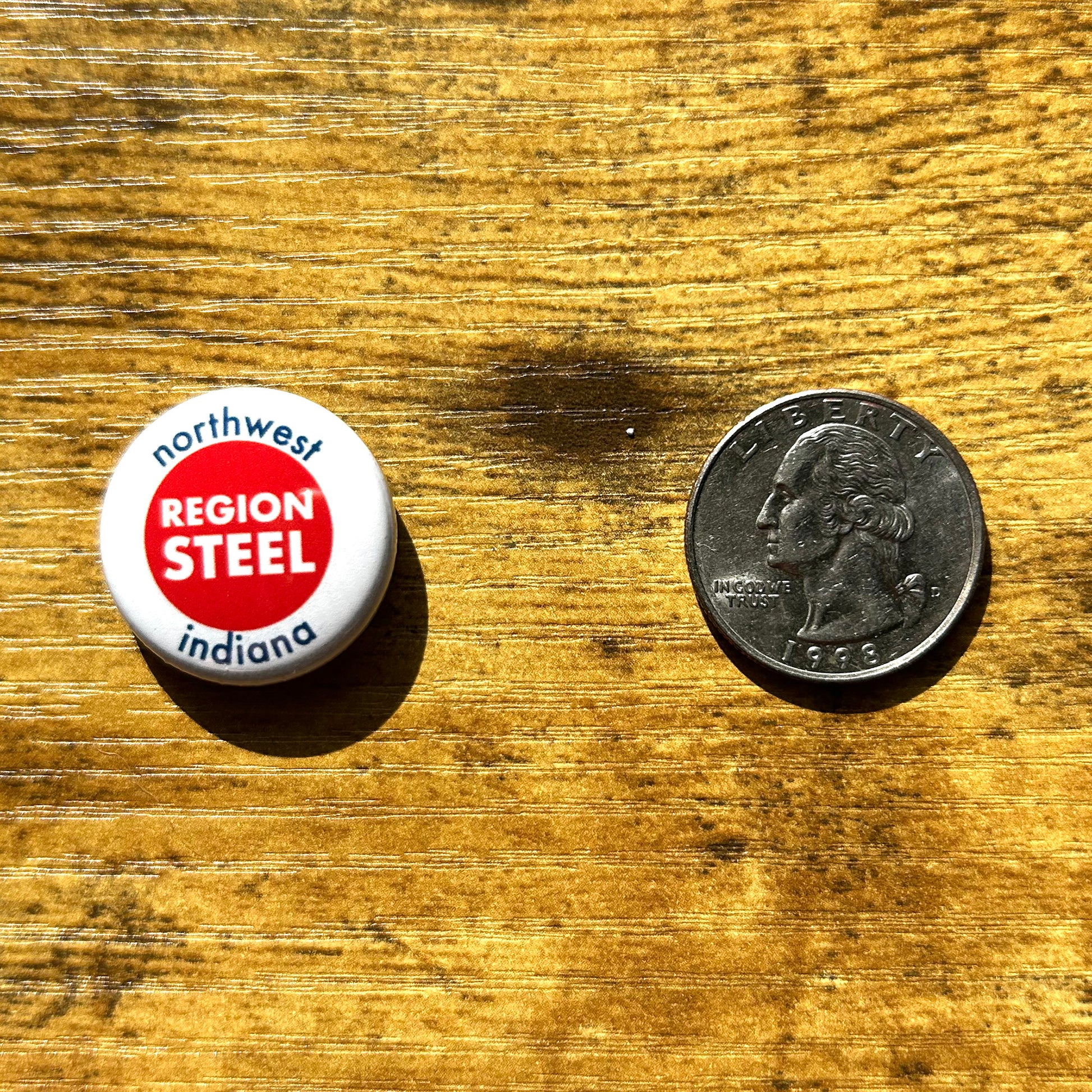 Northwest Indiana Region Steel Pin white pin with red circle in middle says region steel white letters and Northwest Indiana blue letters shown on wood table next to quarter