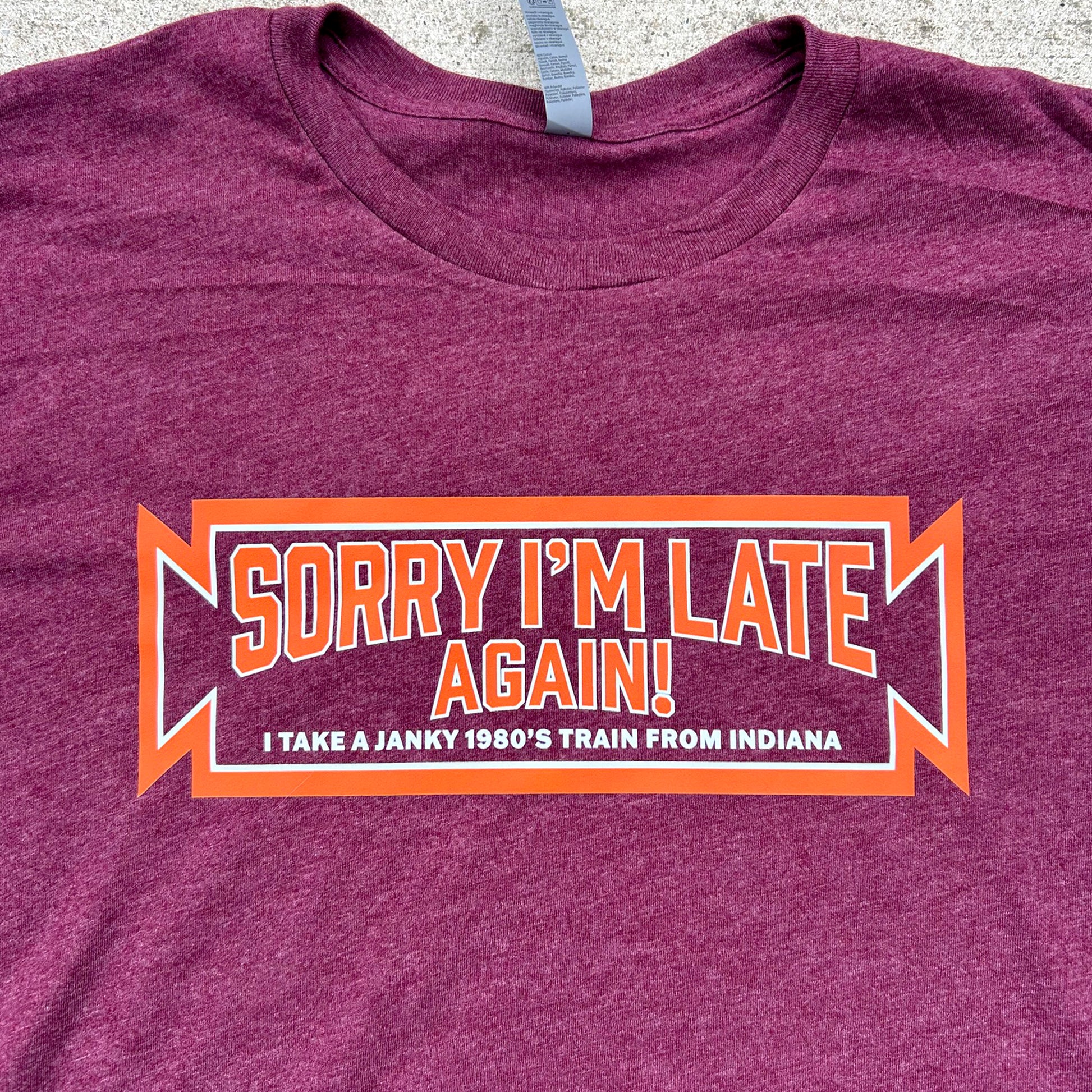 Maroon t-shirt with humorous text design that says "Sorry I'm Late Again! I take aa junky 1980's train from Indiana" on a textured surface